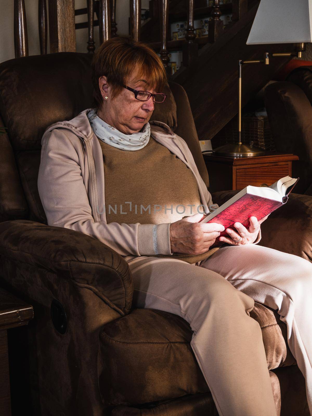 Old woman reading a reed book with house clothes in a sofa. Grandmother reading in house with natural light.