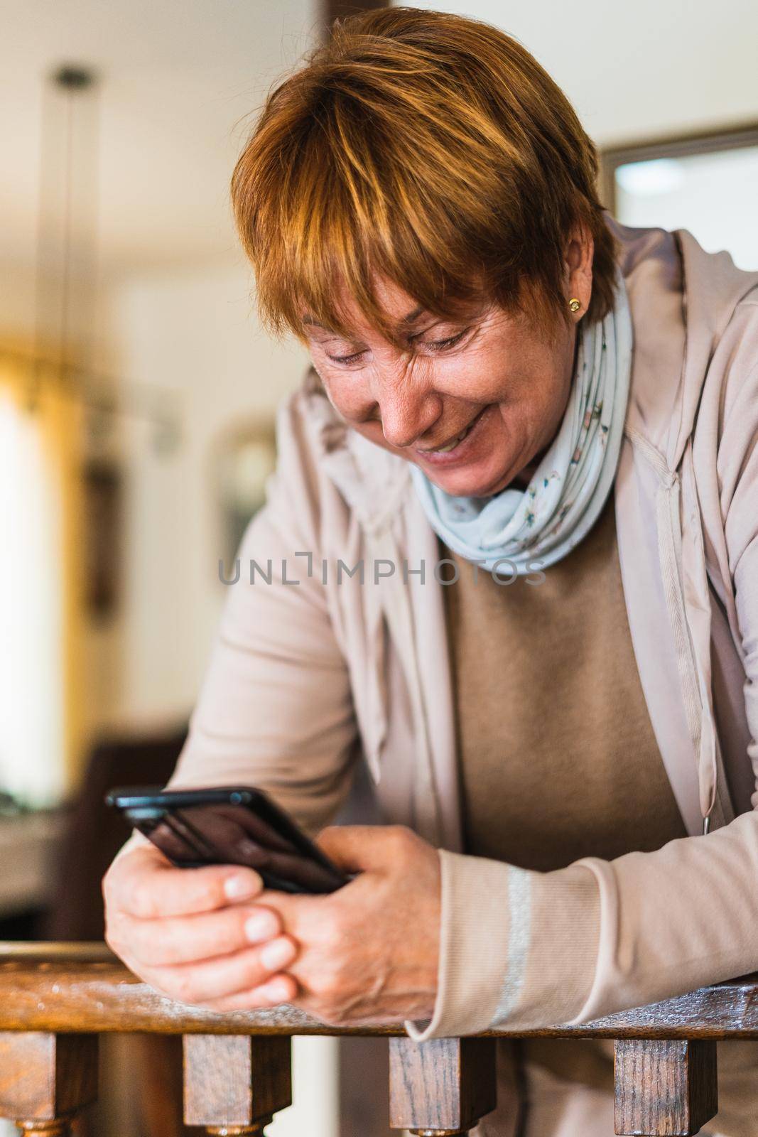 Old woman searching in mobile phone in living room. She wear casual clothes. Caucasian old woman portrait with smartphone. Vertical shot with natural light in home.