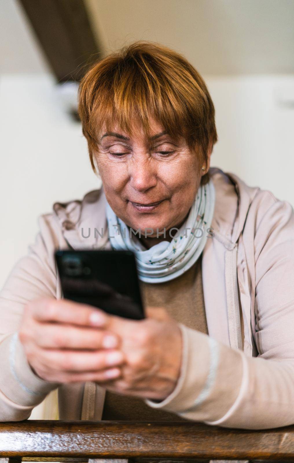 Old woman searching in mobile phone in living room. She wear casual clothes. Caucasian old woman portrait with smartphone. Vertical shot in home.