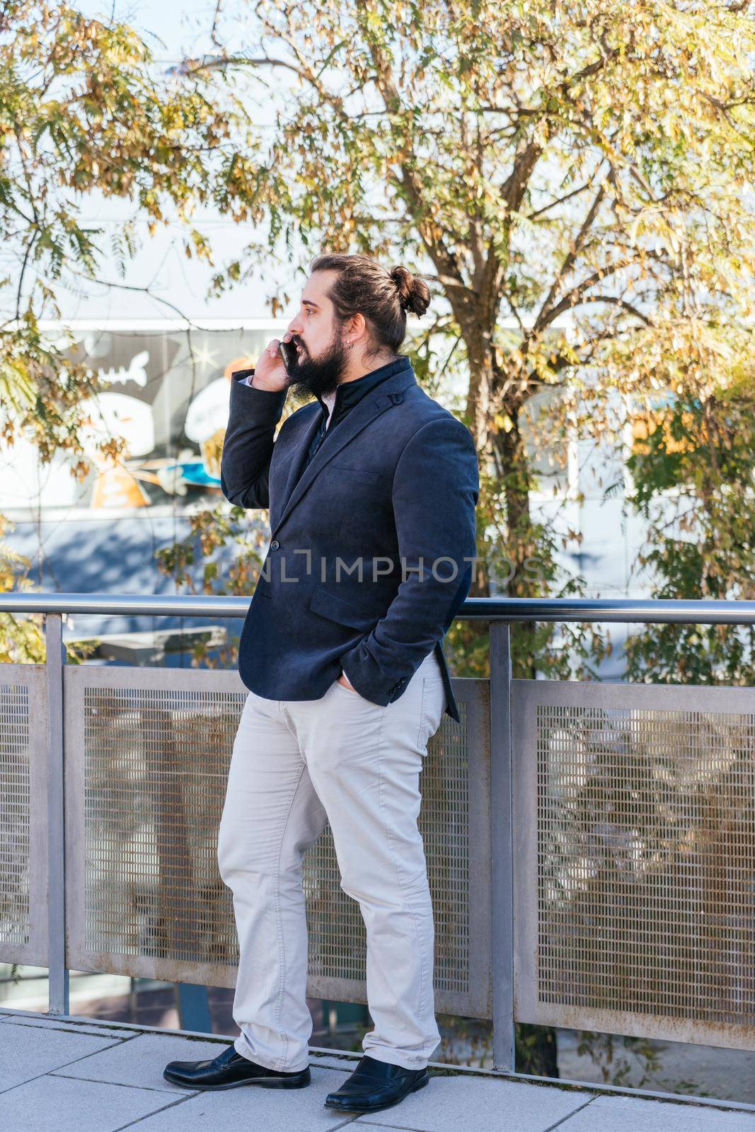 Young businessman with long hair and beard calling during his work break, on the street next to the offices. Vertical photo on a sunny and clear day.
