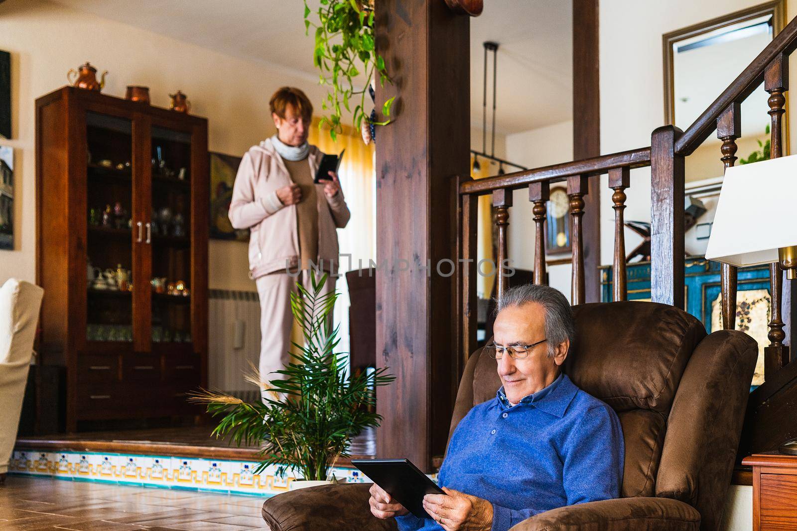 Grandfather using tablet in a living room with natural light. He wear a blue sweater and glasses. Its in his house, size view with natural light