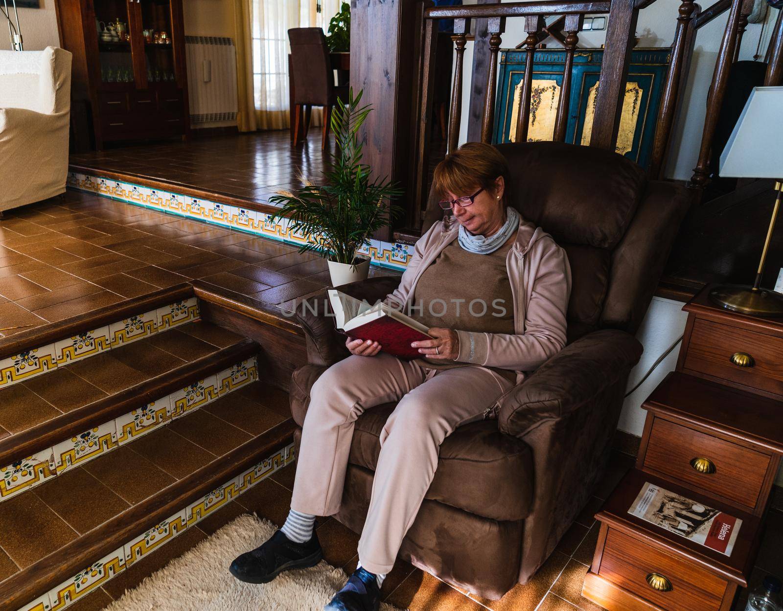 Angular view of a grandmother reading a red book with purple glasses. She is relaxed in her home. Its a rustic home with natural light