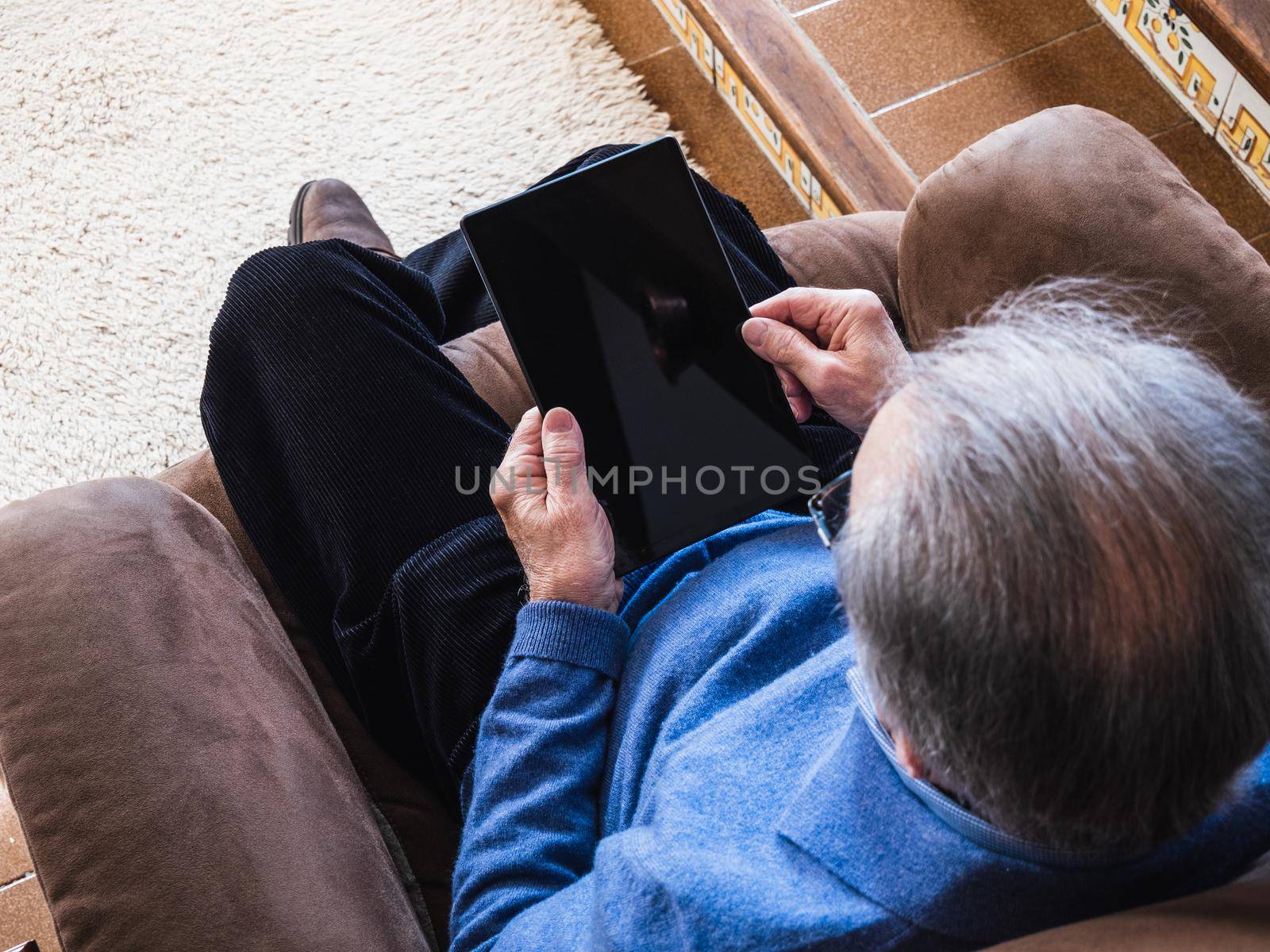 Grandfather man looking a tablet in living room. He is in a sofa and wear a blue sweater. He have glasses. Natural light in house
