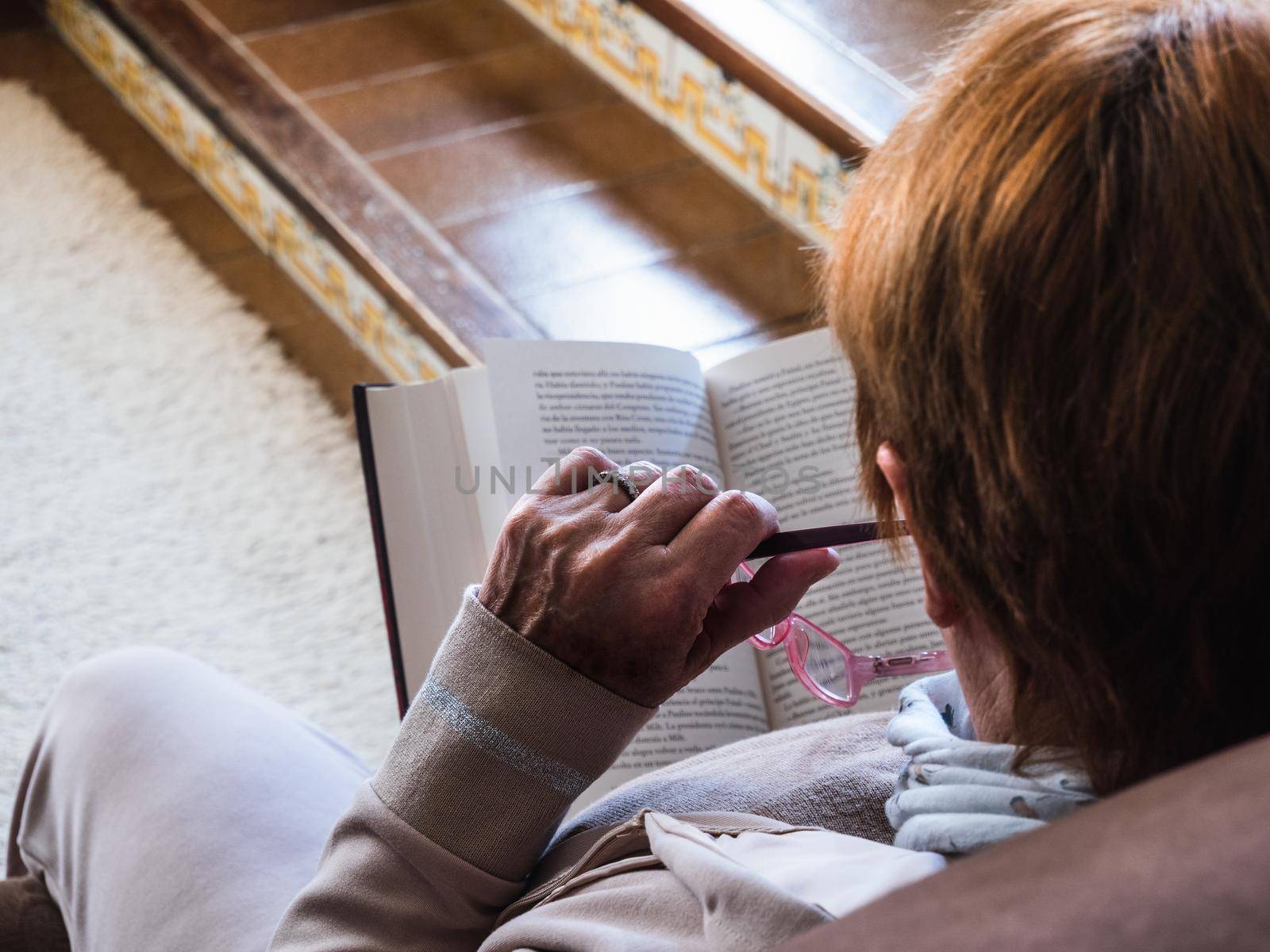 Detail back view of caucasian woman reading a book with a purple glasses in her hand. She is in her living room with natural light