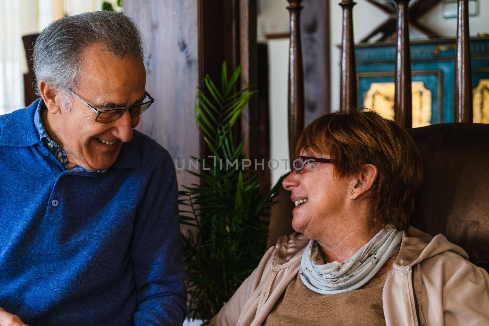 Old couple looking at each other with love in living room. She have purple glasses and is sited in a sofa and he have a silver glasses and blue sweater. They are in a living room with natural light