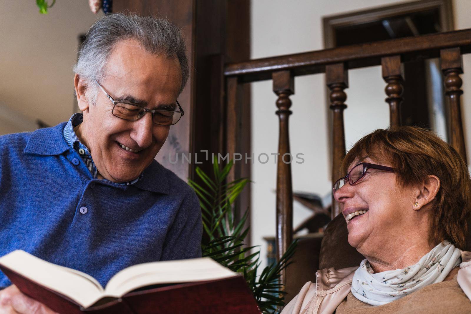 Old marriage reading a red book sited in a sofa in living room. He wear a blue sweater and glasses. She have a purple glasses and its smiling looking to her husband. They are in house with natural light.