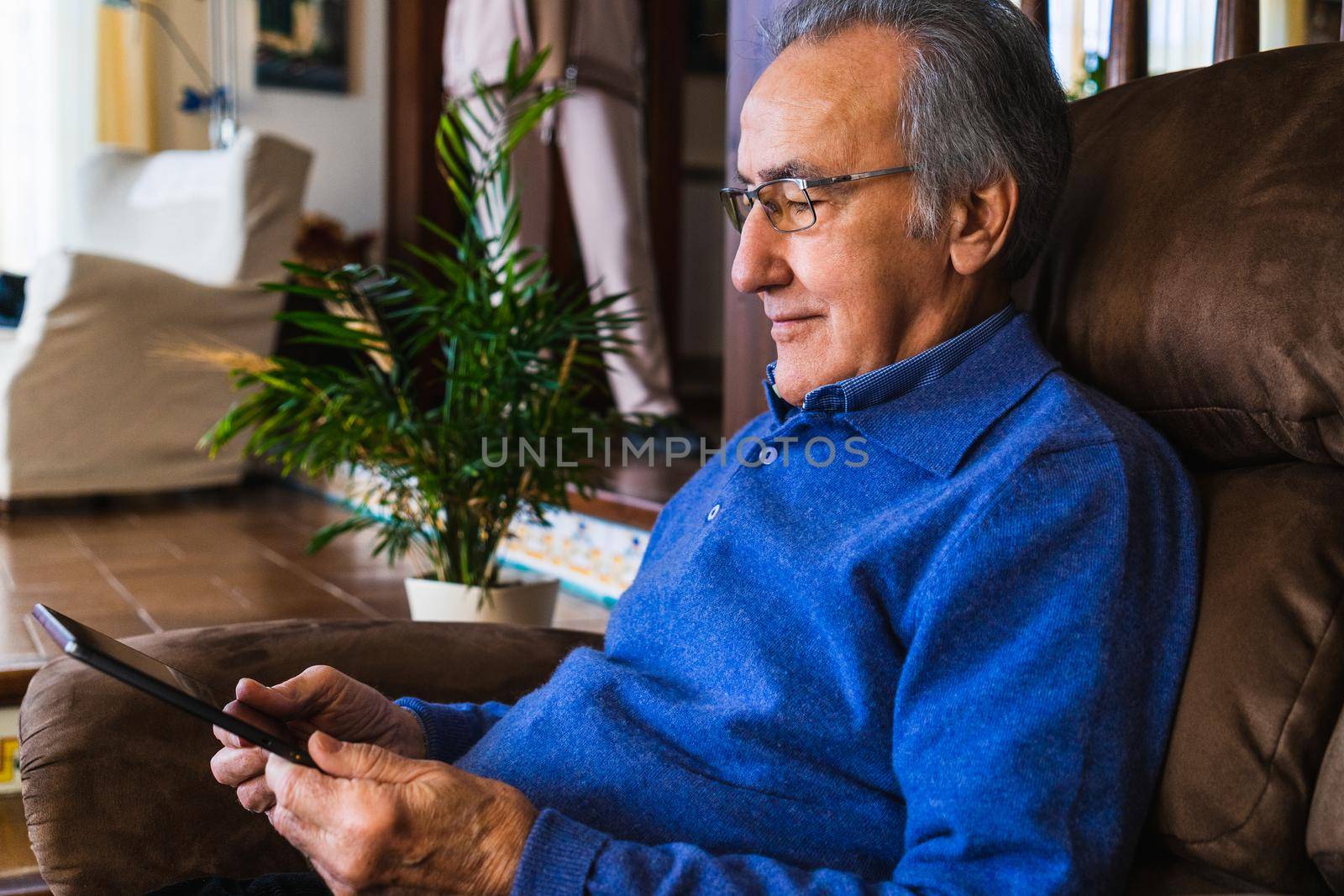 Grandfather using tablet in a living room with natural light. He wear a blue sweater and glasses. Its in his house, size view with natural light