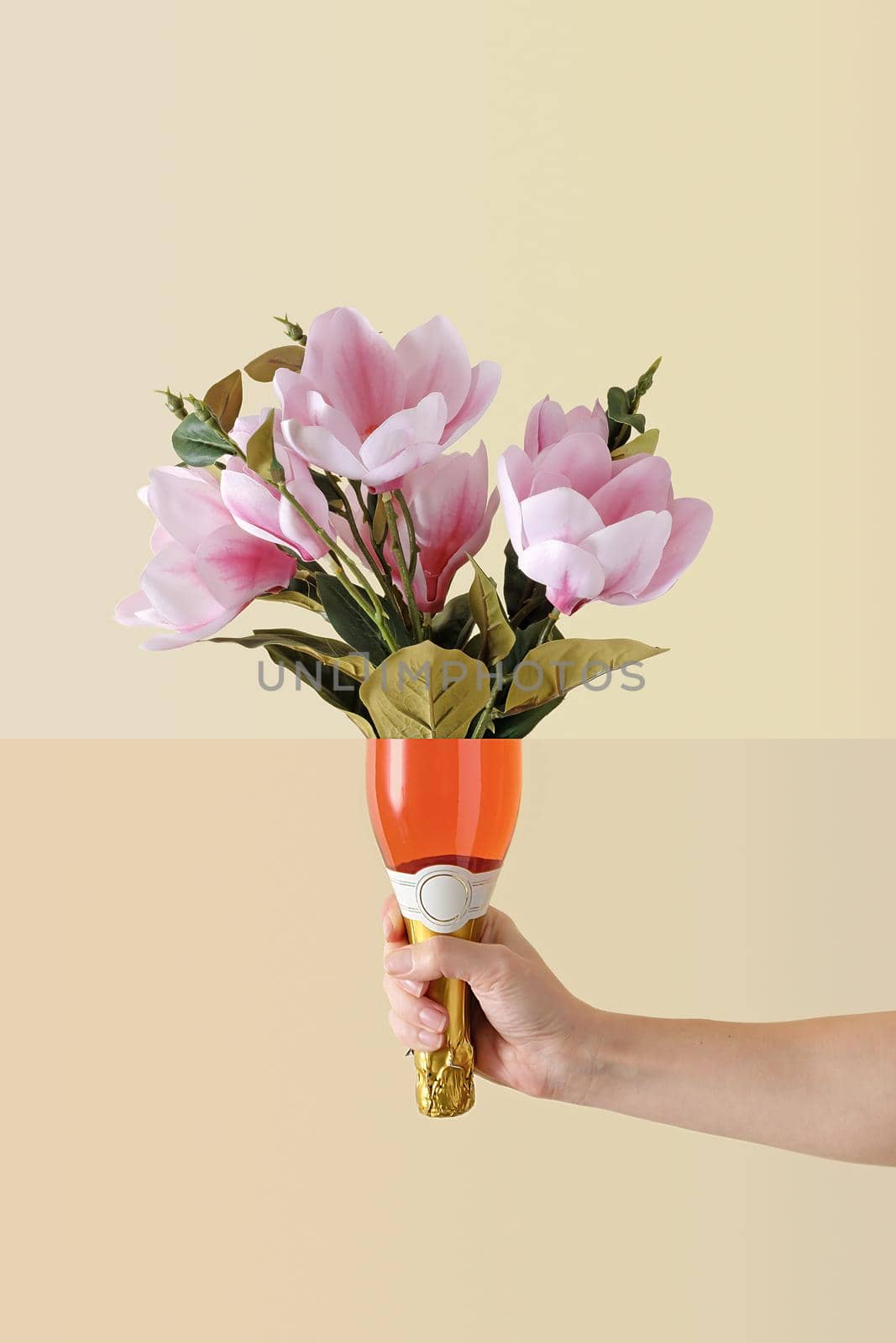 Conceptual image of fresh summer flowers in a luxury rose champagne bottle held by an extended hand over a pale yellow background for a special occasion