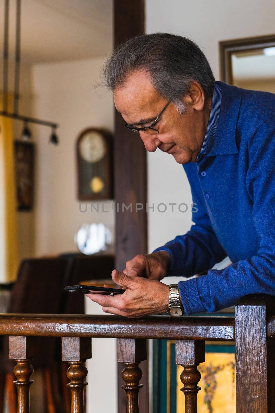 Old man looking smartphone stand up in home. He wear a blue sweater and glasses. Vertical shot of caucasian senior male with phone with natural light.