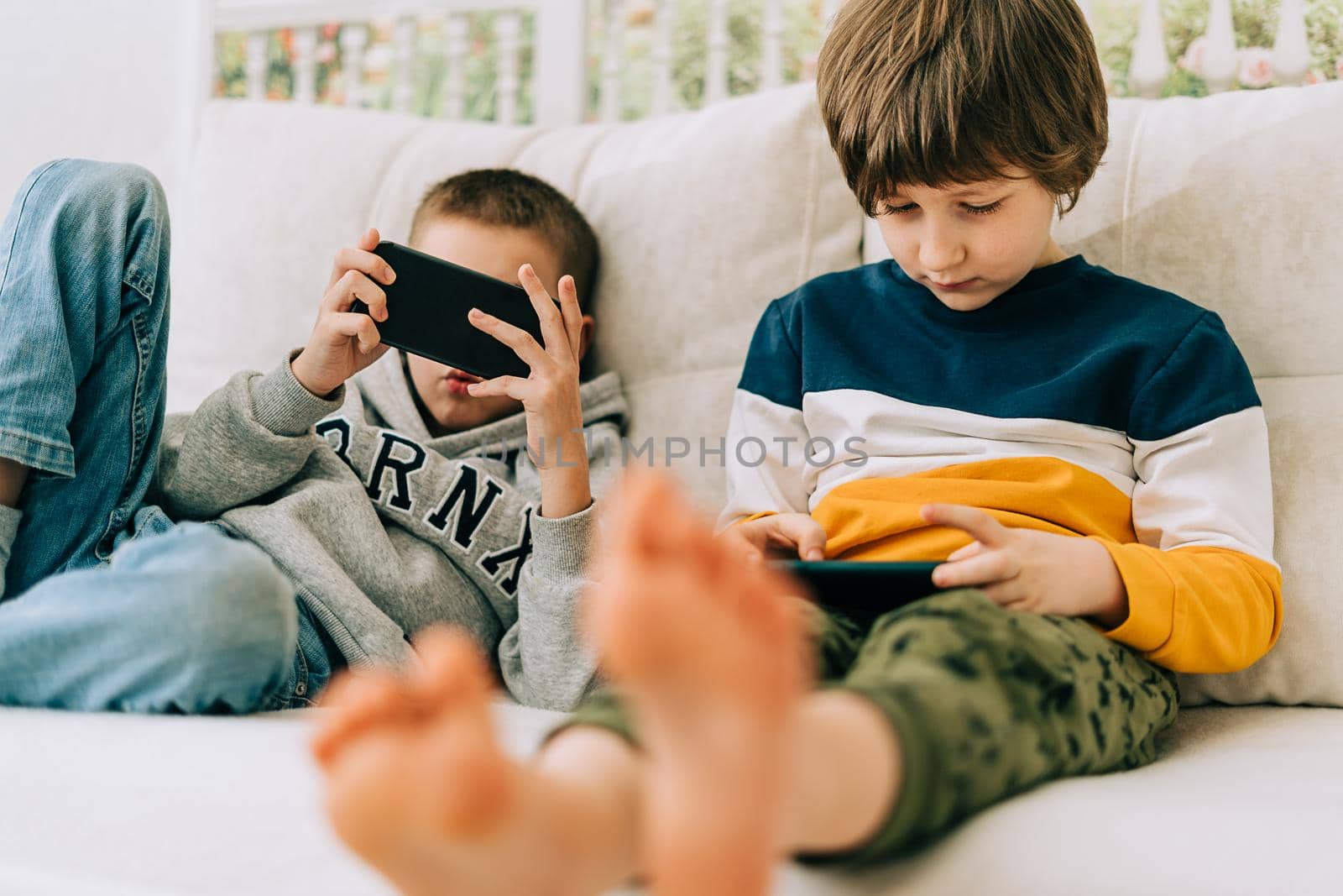 Cheerful two brothers boys playing online game, watching video on cellphone. Smiling kids using funny mobile apps, enjoying free leisure time at home on couch. Children texting together at phone.