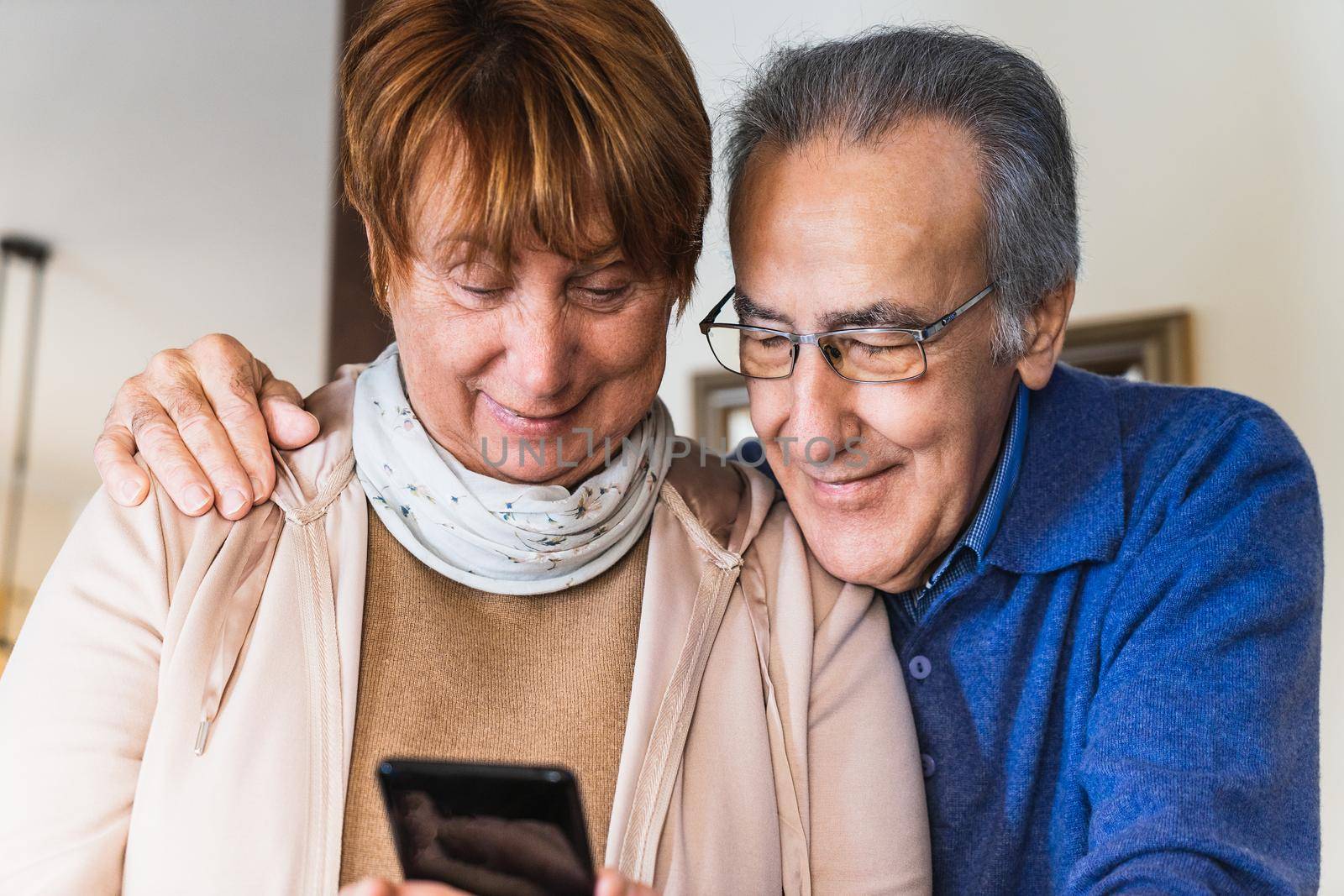 Grandparents looking smartphone in living room with natural light. Marriage together in home. She have the phone and he wear blue sweater and glasses. They are stand up in a rustic and traditional house