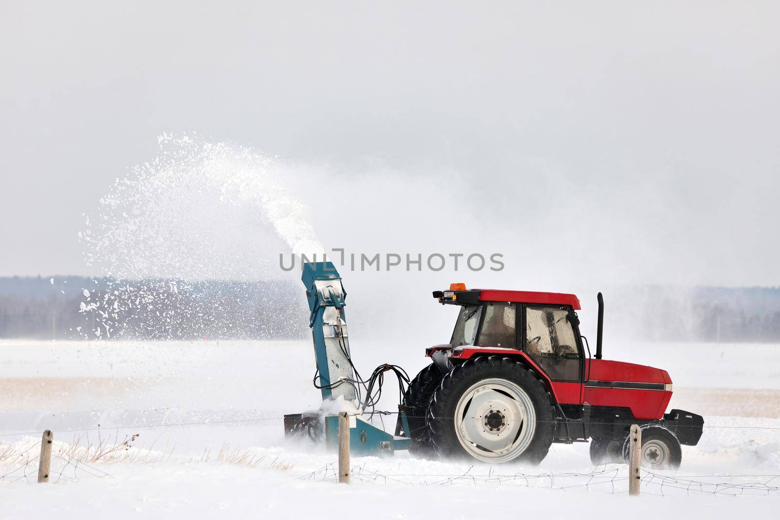 Red Tractor with snowblower attachment Snow Blowing a Driveway in a Rural Setting. It's windy and the blown snow creates a large plume of snow. High quality photo