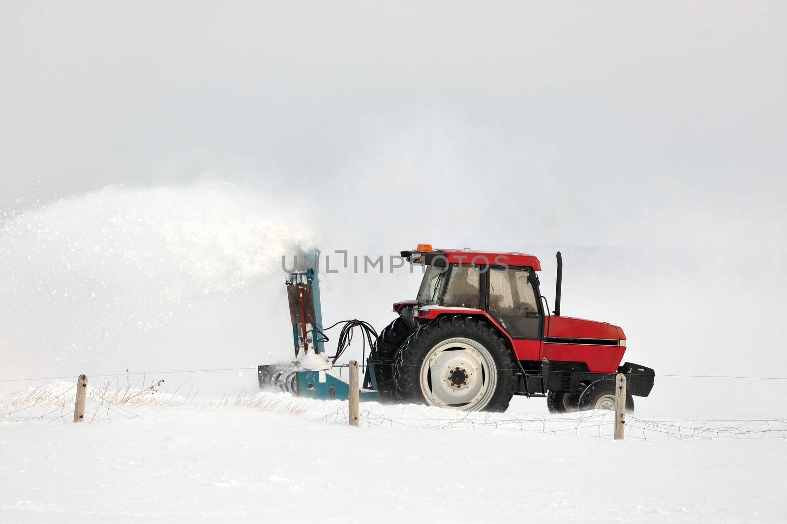 Red Tractor with snowblower attachment Snow Blowing a Driveway in a Rural Setting. It's windy and the blown snow creates a large plume of snow. High quality photo