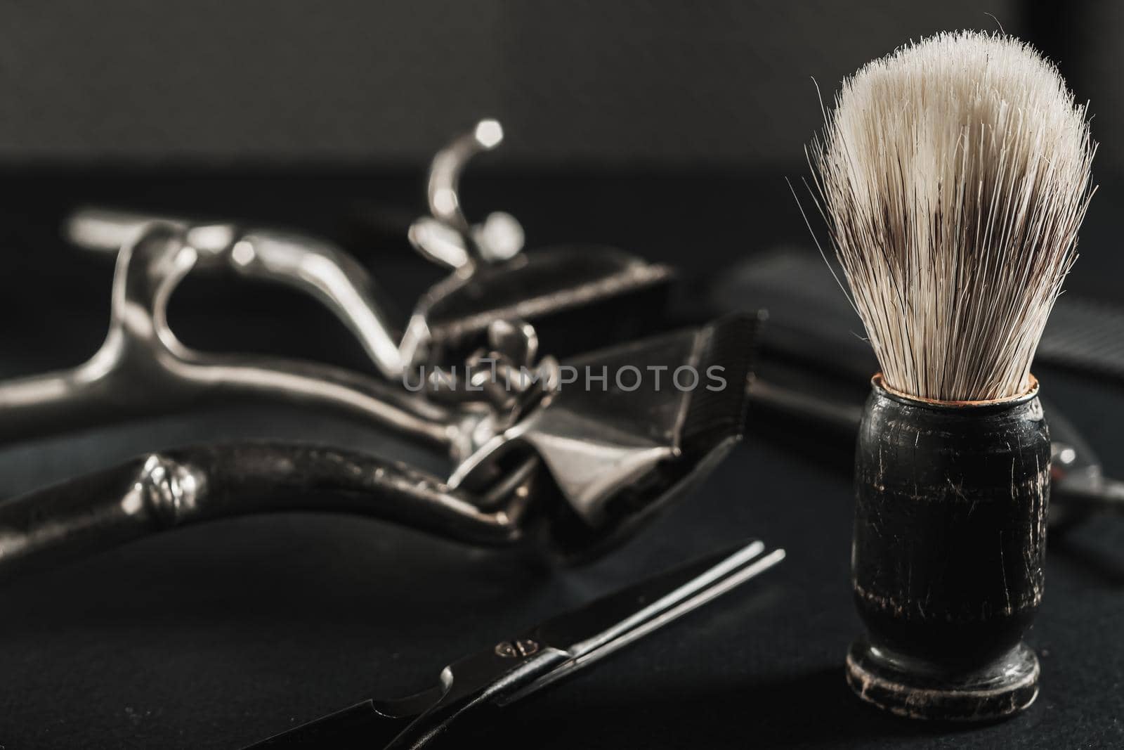 On a black surface are old barber tools. shaving brush vintage manual hair clipper, comb, razor, hairdressing scissors. black monochrome. Close-up. Barbershop background. contrast shadows. Horizontal
