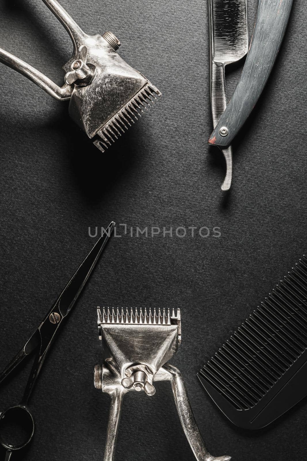 On a black surface are old barber tools. two vintage manual hair clipper, comb, razor, hairdressing scissors. black monochrome. Close-up. Barbershop background. contrast shadows. vertical orientation. Top view, flat lay.