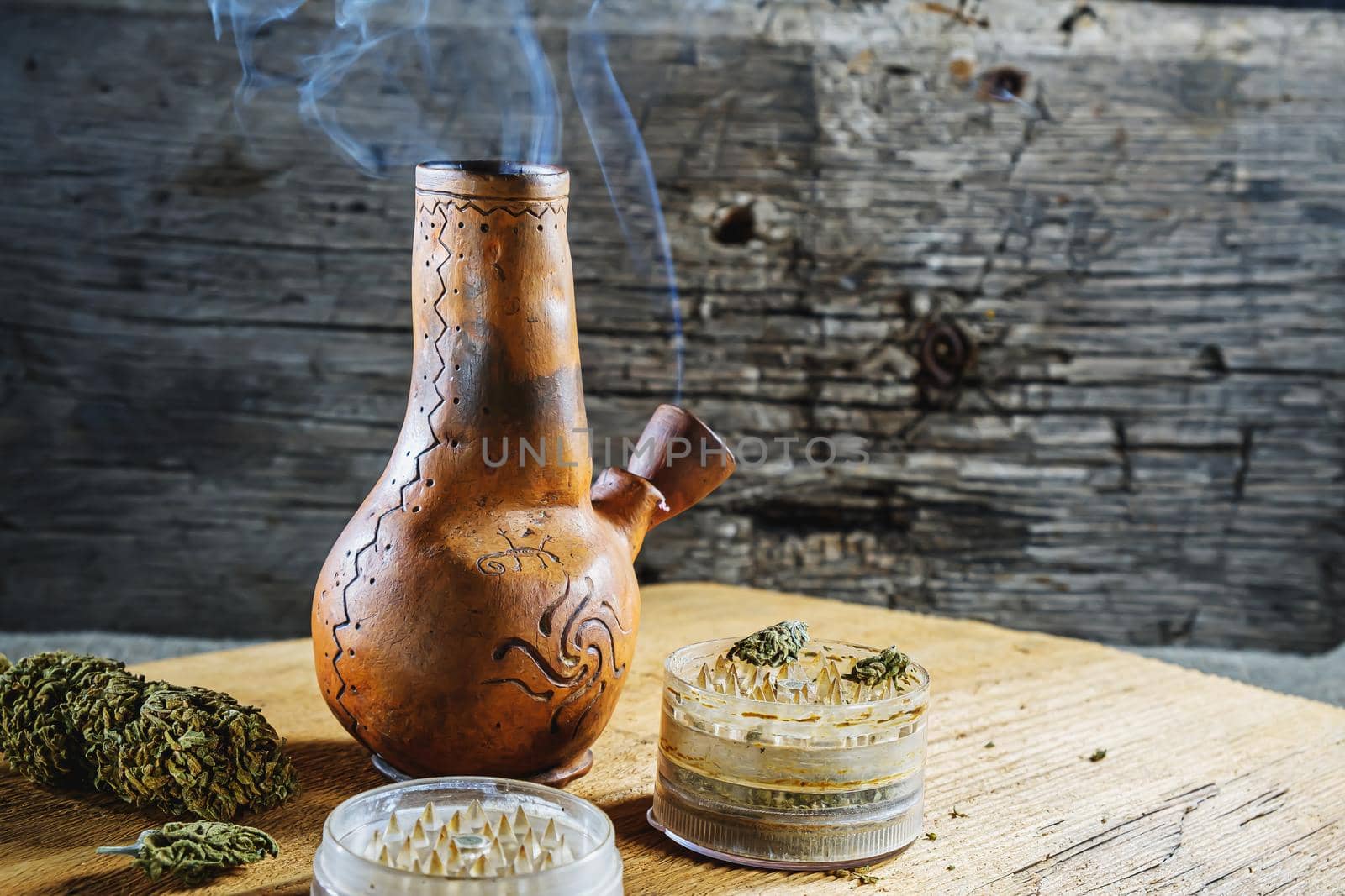 Old brown steamung bong and open cannabis grinder with chopped marijuana weed and medical cannabis bud in smoke by wooden desck and sackcloth. Horizontal orientation