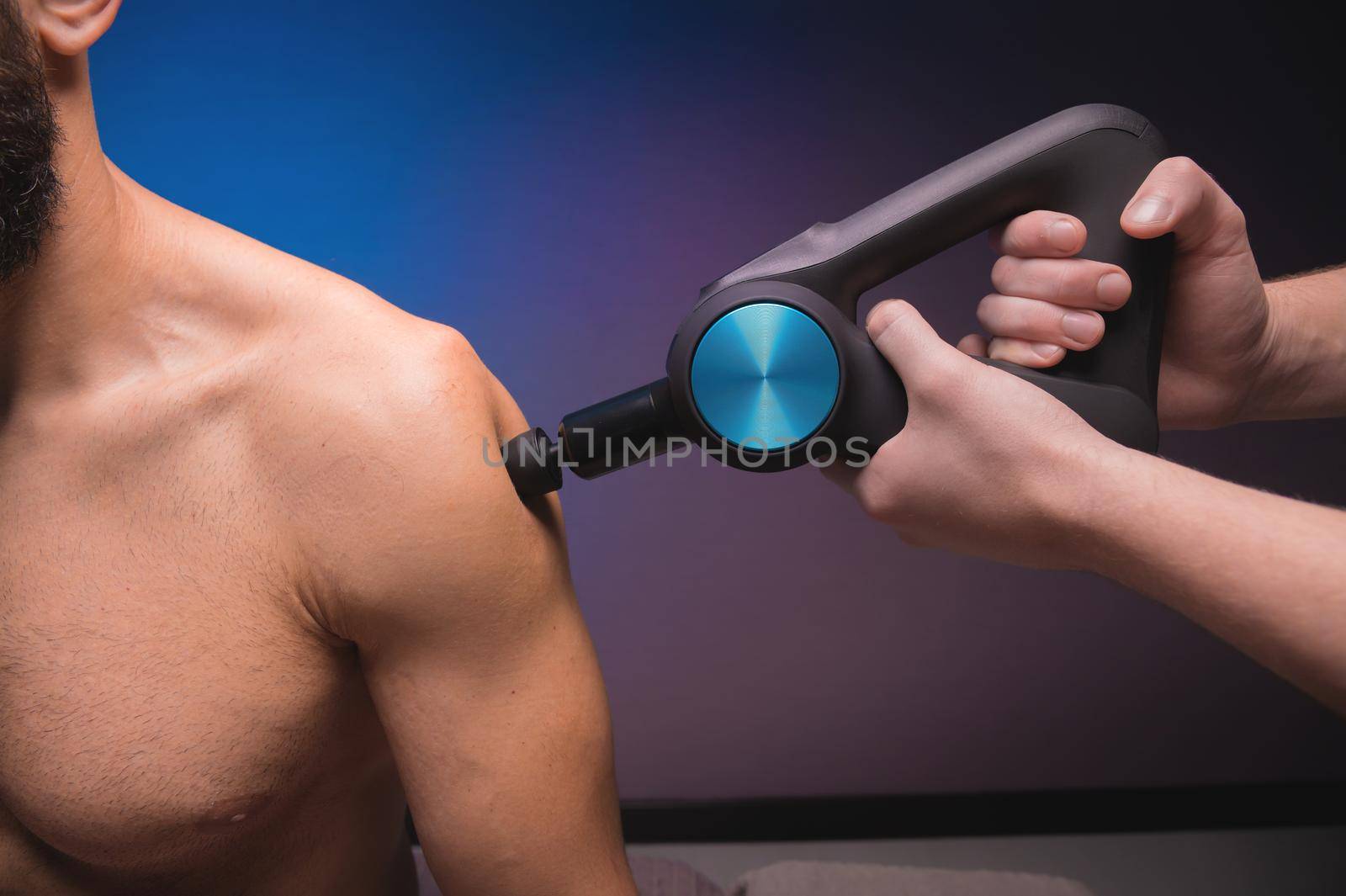 Close-up percussion massager in the hands of a masseur makes a massage of the shoulders and upper back to a male client lying down.