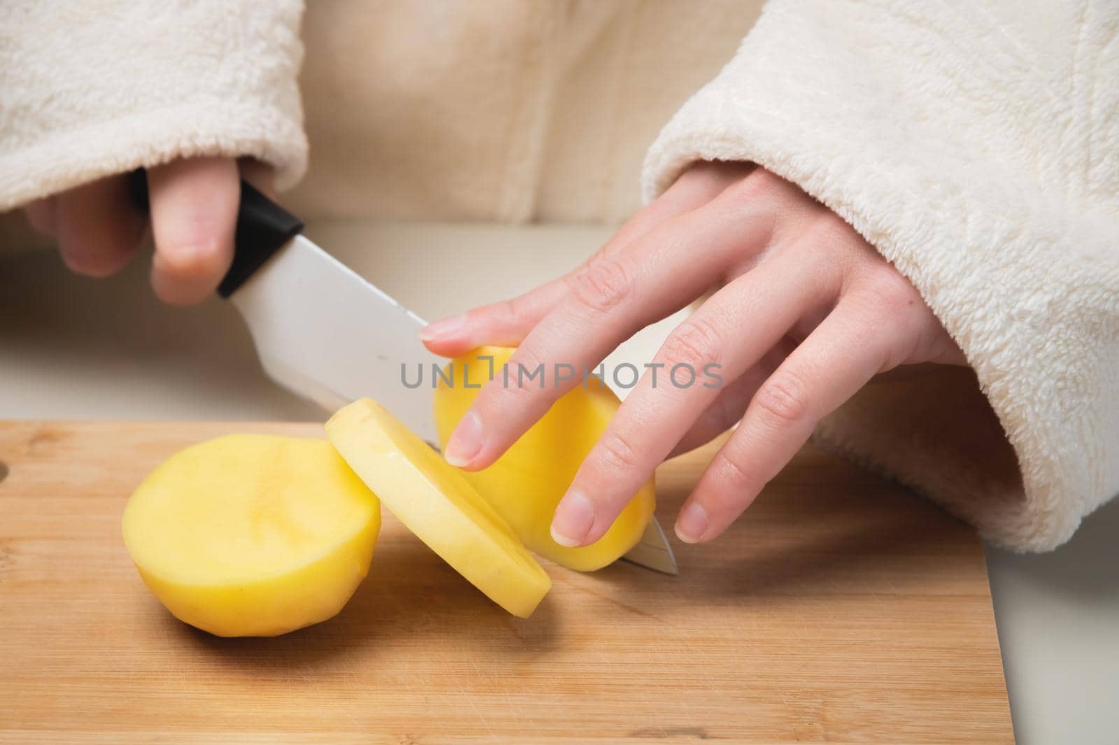 young caucasian woman in a bathrobe stands in the kitchen and cuts fresh potatoes with a knife, on a wooden cutting board