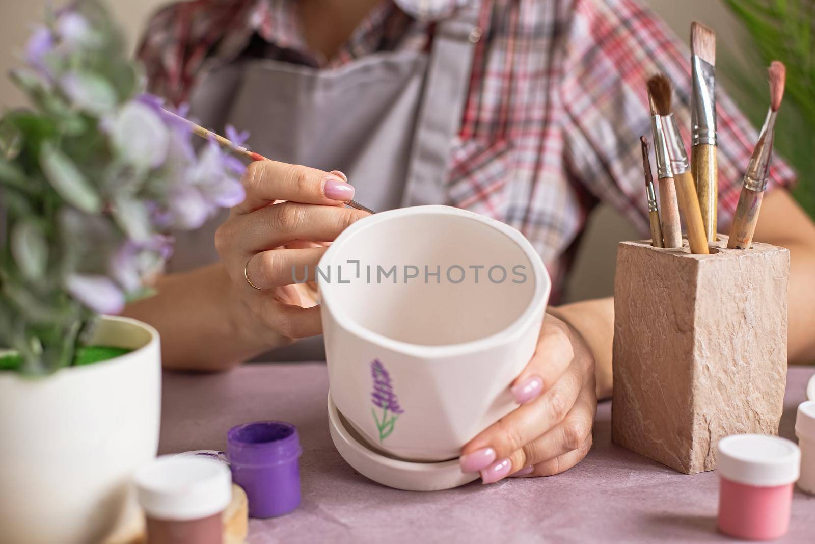 A hands of woman in a gray apron paints a white ceramic flower pot at the table in daylight. On the table is purple kraft paper, brushes, jars of paints. Close-up. No face