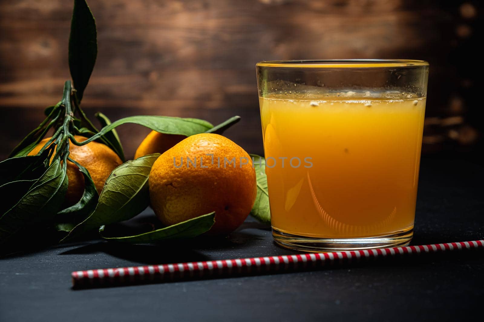on a dark surface are three tangerines with leaves next to a glass of juice. Nearby lies a red and white striped straw. Horizontal orientation.