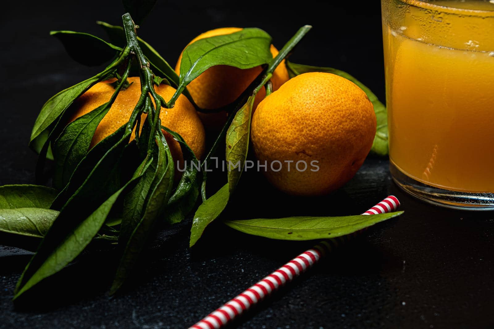 on a dark surface are three tangerines with leaves next to a glass of juice. Nearby lies a red and white striped straw. Horizontal orientation.