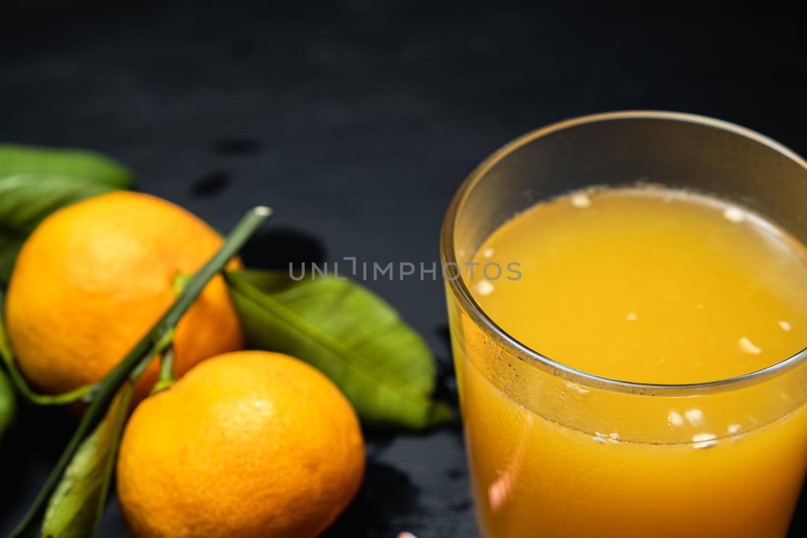 on a dark surface are three tangerines with leaves next to a glass of juice. Horizontal orientation.