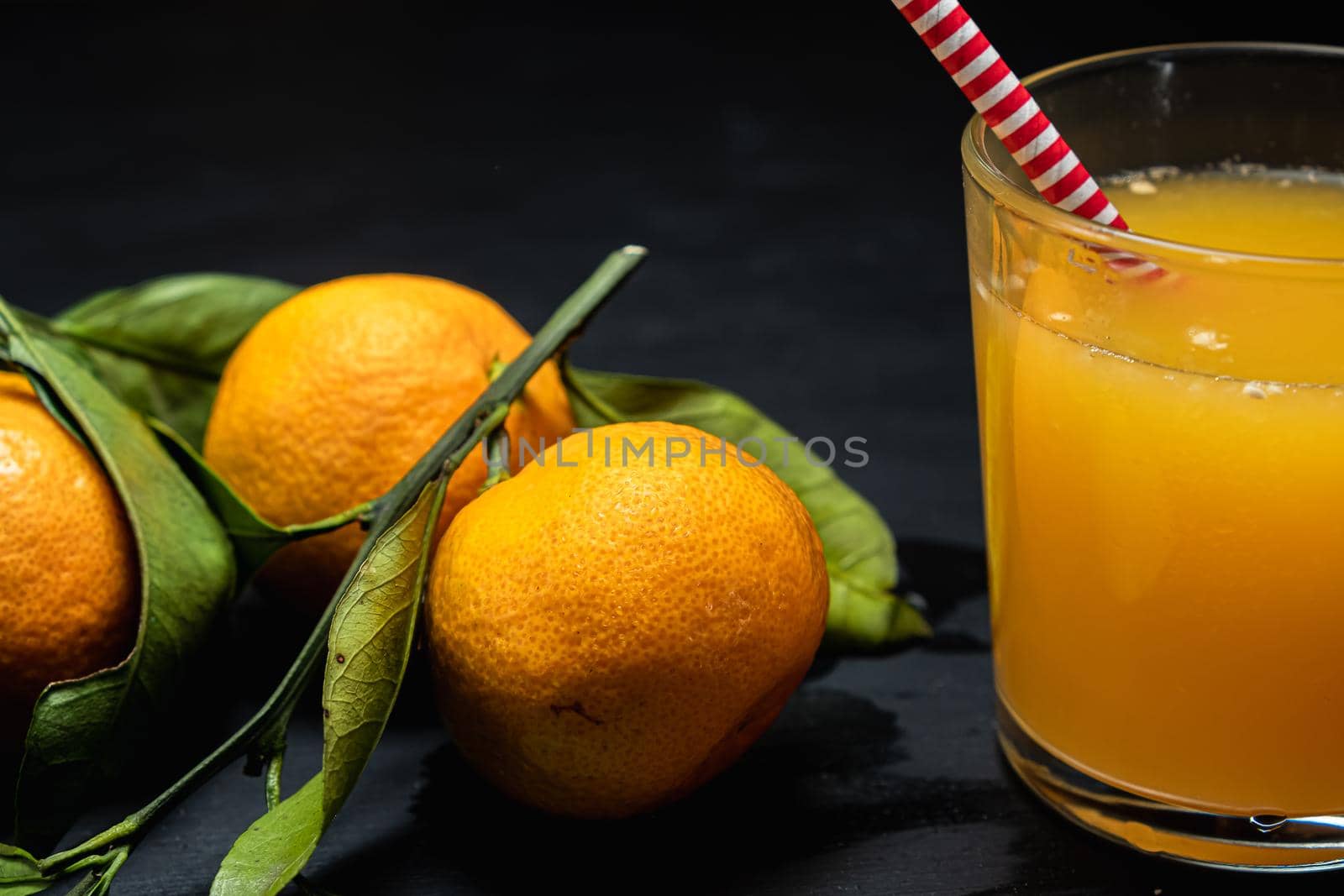 on a dark surface are three tangerines with leaves next to a glass of juice. Horizontal orientation.