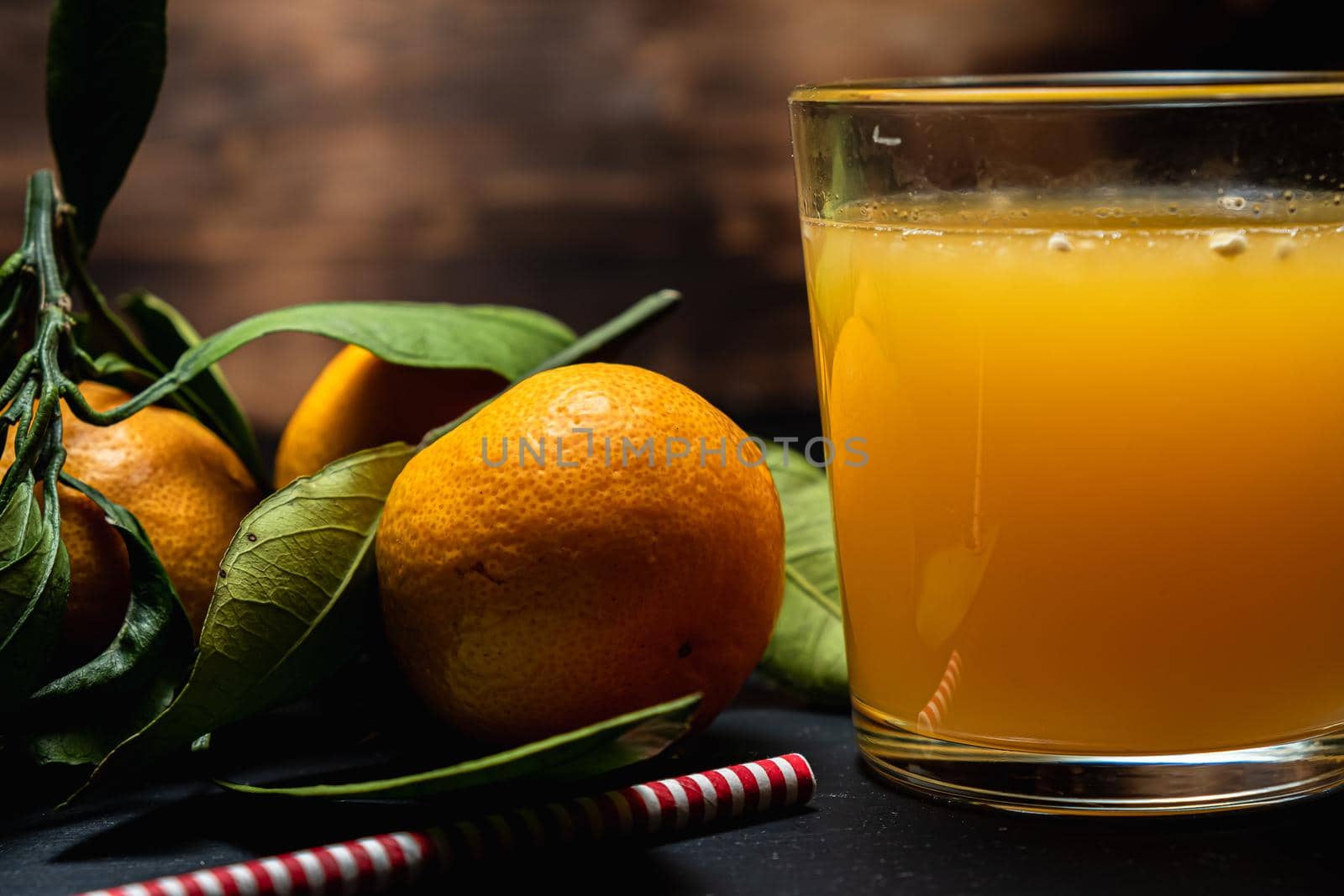 on a dark surface are three tangerines with leaves next to a glass of juice. Nearby lies a red and white striped straw. Horizontal orientation.