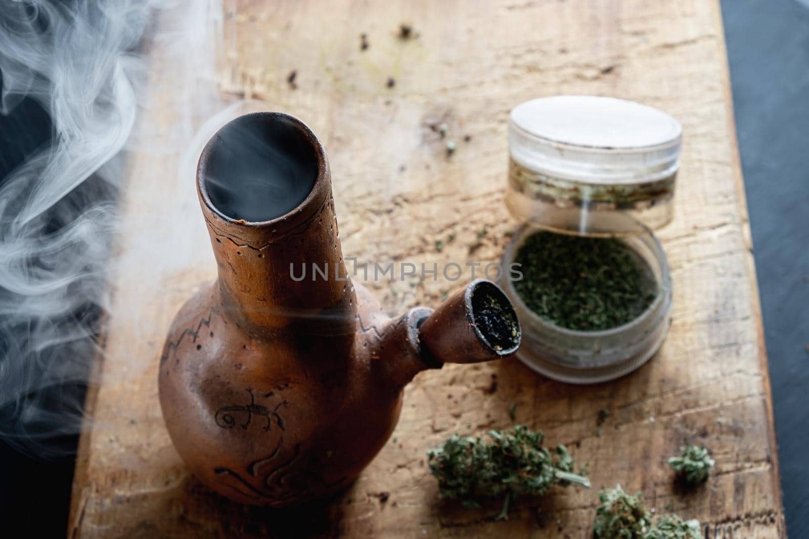 Old bong and open cannabis grinder with chopped weed and marijuana in smoke are located on the board. Wooden background. Horizontal composition.