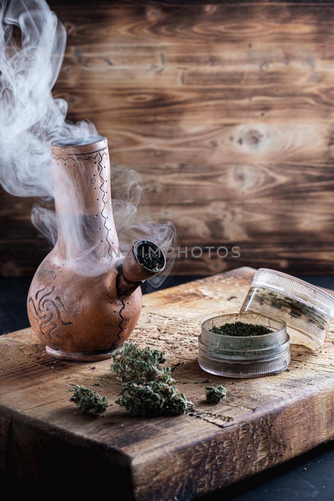 Old bong and open grinder cannabis and marijuana in smoke are located on the board. Wood background. Vertical composition.