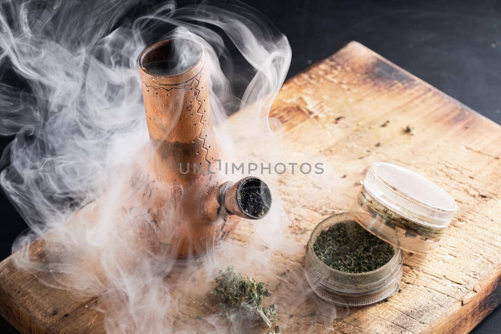 Old bong and open grinder cannabis and marijuana in smoke are located on the board. Wood background. Horizontal composition.