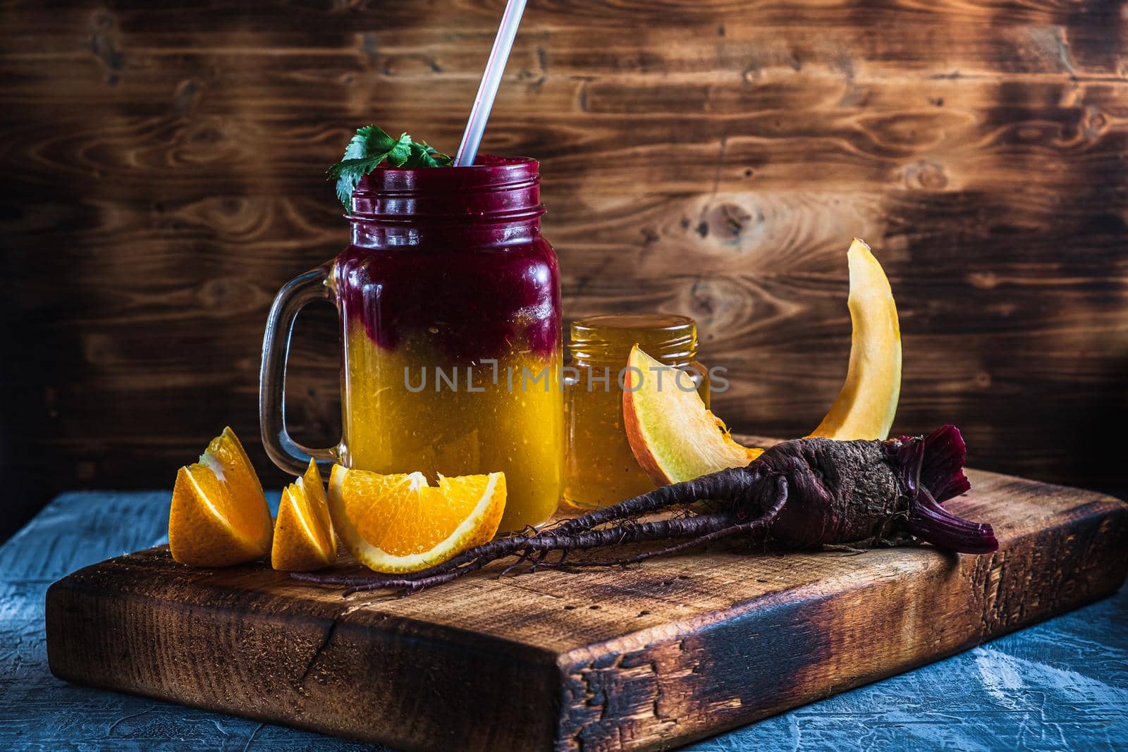 smoothie consists of beetroot pumpkin, orange, celery in mason jar. smoothies are poured in layers. next to it is a jar of honey, beet root, a piece of pumpkin and slices of orange. on a thick wooden board. horizontal orientation.