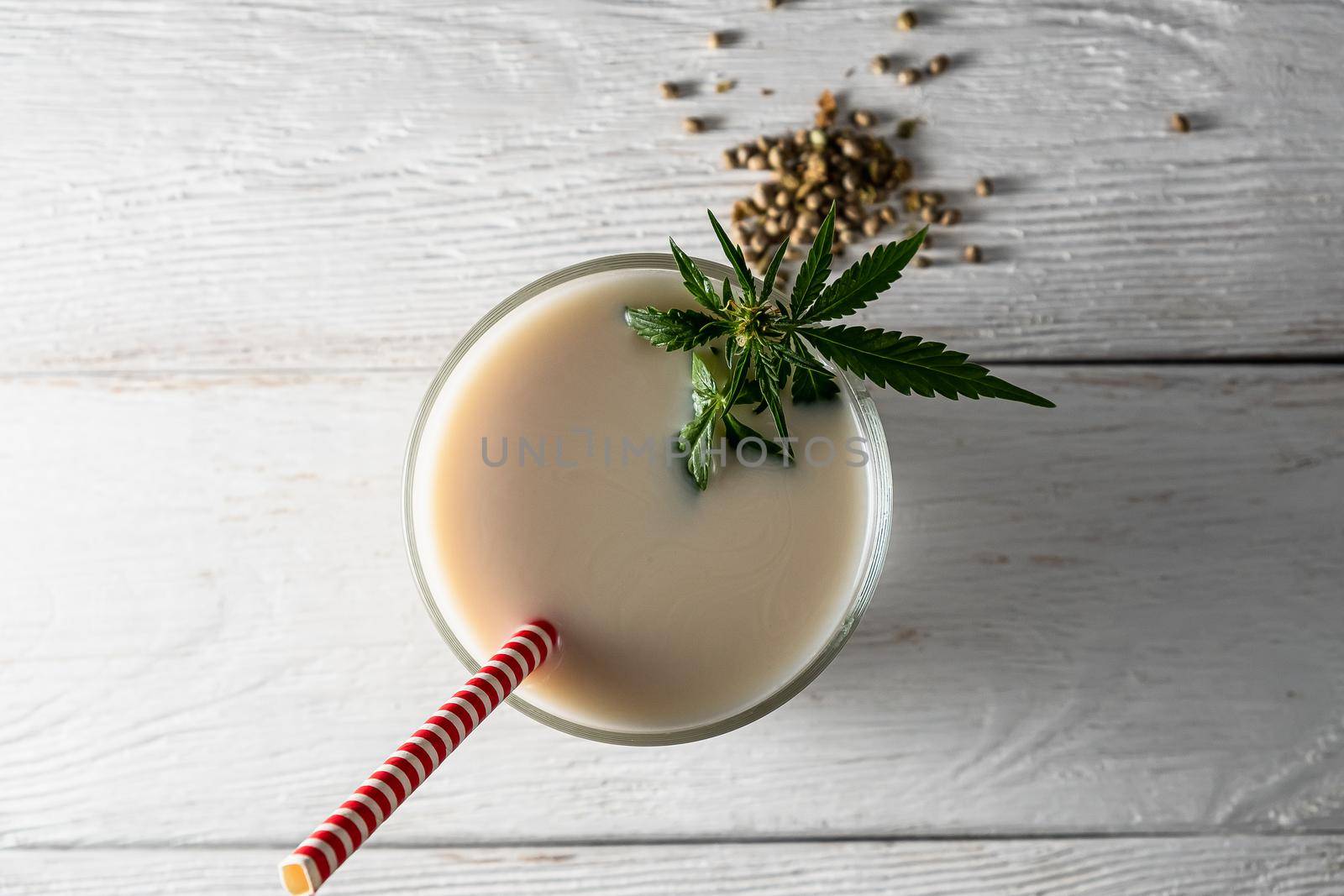 Hemp milk is poured into a glass. On the surface is a cannabis leaf. Nearby marijuana seeds. On the white old weathered boards. In a glass, red and white non-plastic straw. flat lay top view