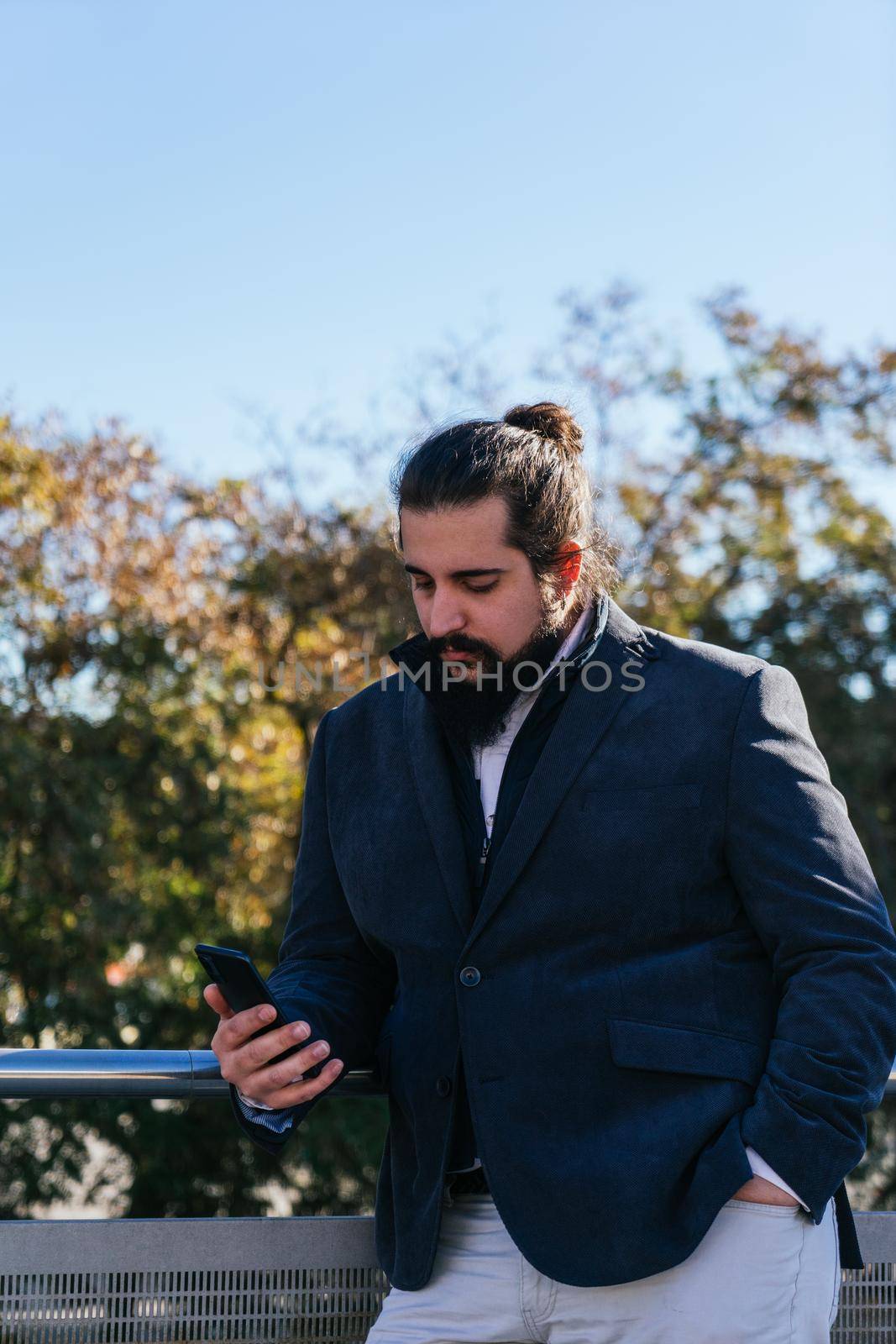 Young businessman with long hair and beard looking smartphone during his work break, on the street next to the offices. Vertical photo on a sunny and clear day.