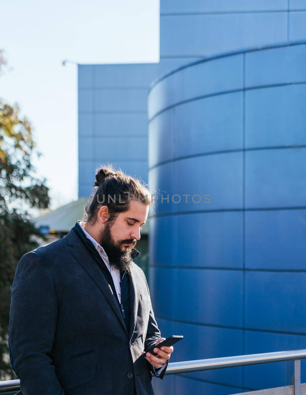 Young businessman with long hair and beard looking smartphone during his work break, on the street next to the offices. Vertical photo on a sunny and clear day.