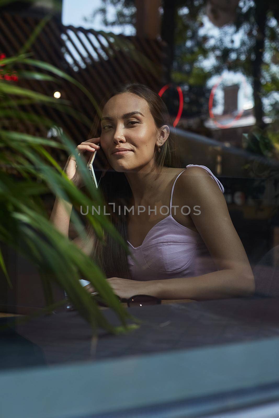 young asian pretty woman sitting at table of coffee shop, calling by smartphone. beautiful smiling lady using cell phone shot through window glass outside of cafe. modern communication technology