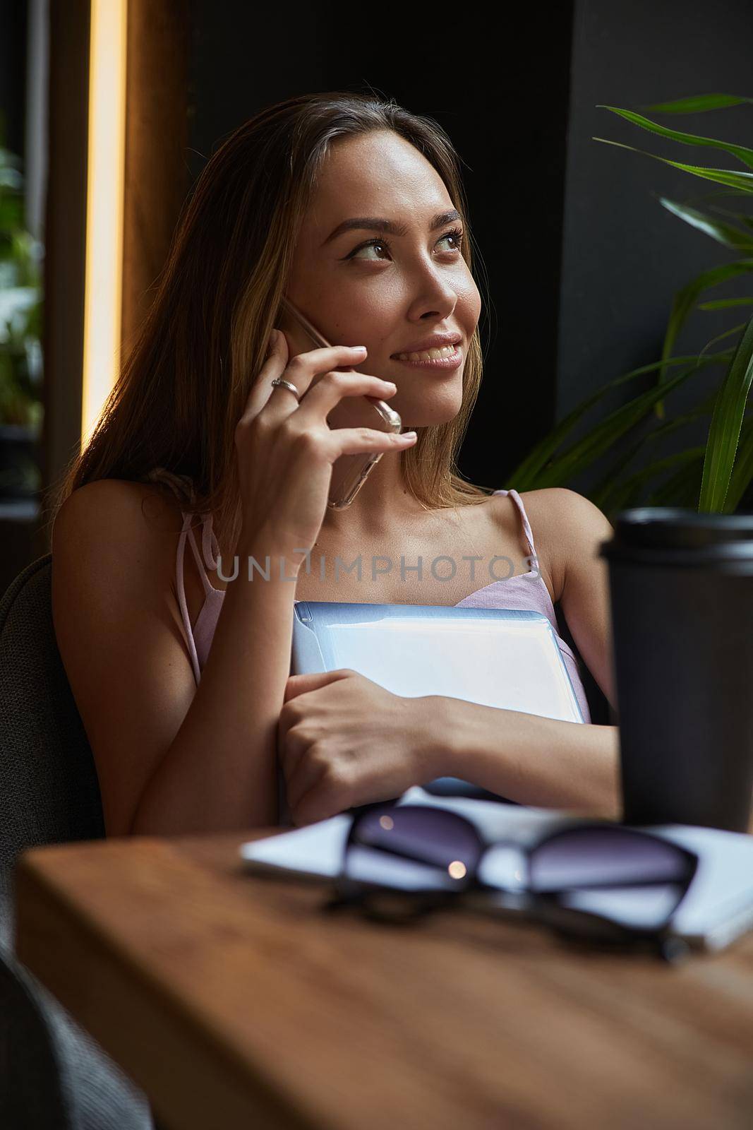 young asian pretty woman sitting at table of coffee shop, calling by smartphone by artemzatsepilin