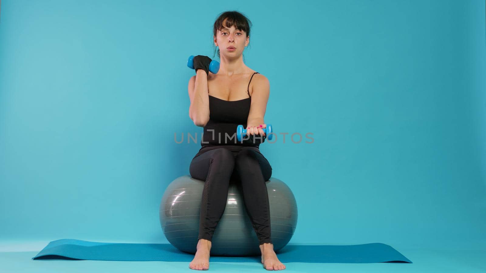 Fit person using dumbbells to lift weights on fitness toning ball, doing physical exercise to train body muscles. Athletic woman using pilates equipment on yoga mat in front of camera.