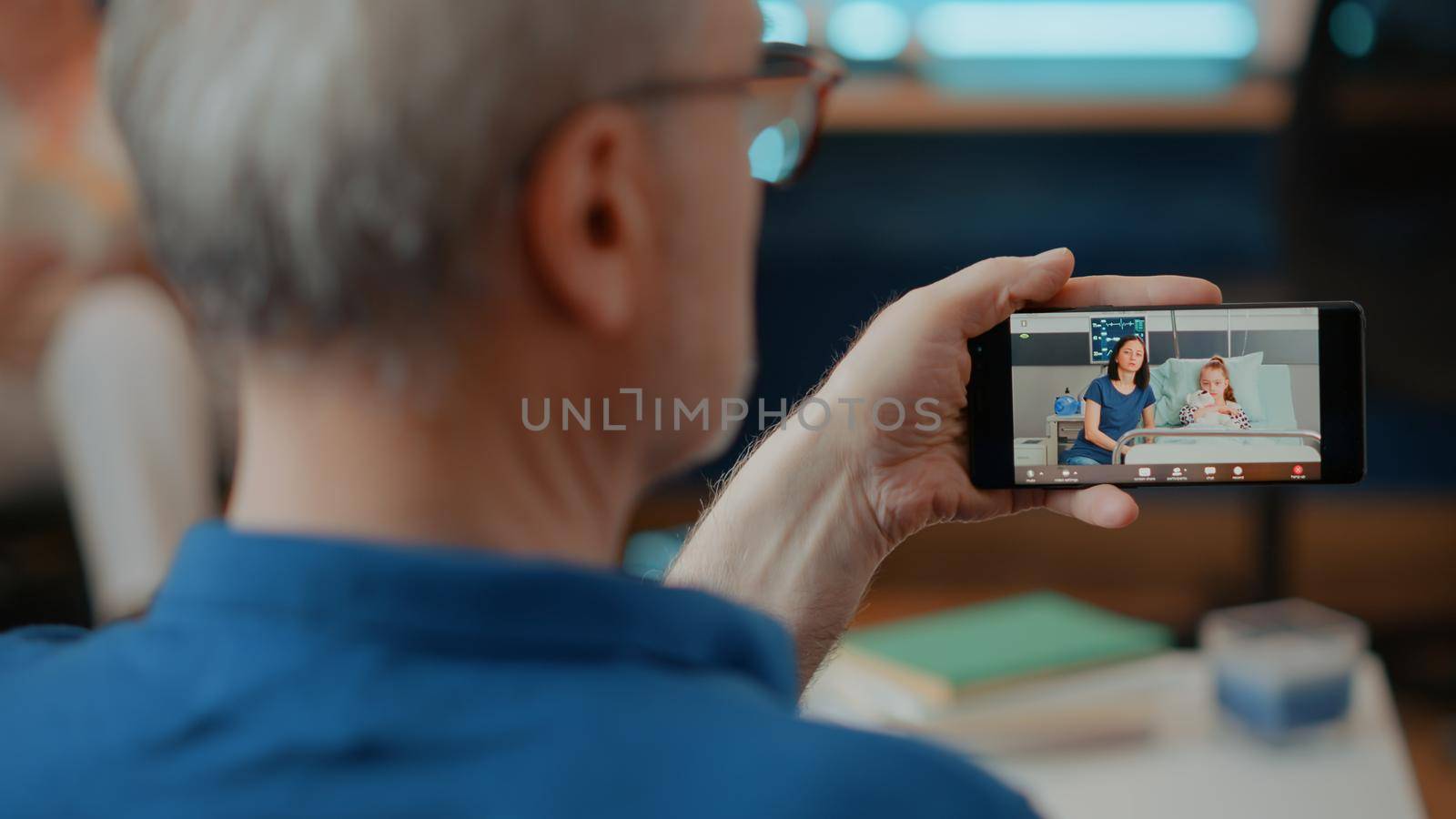 Retired adult using online video call on mobile phone, talking to relatives in hospital. Older man holding smartphone with video teleconference for remote communication with family.