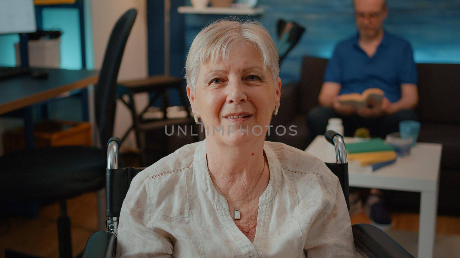 Portrait of retired woman sitting in wheelchair at home. Mature person with chronic disability looking at camera and smiling in living room, receiving transportation support and aid.