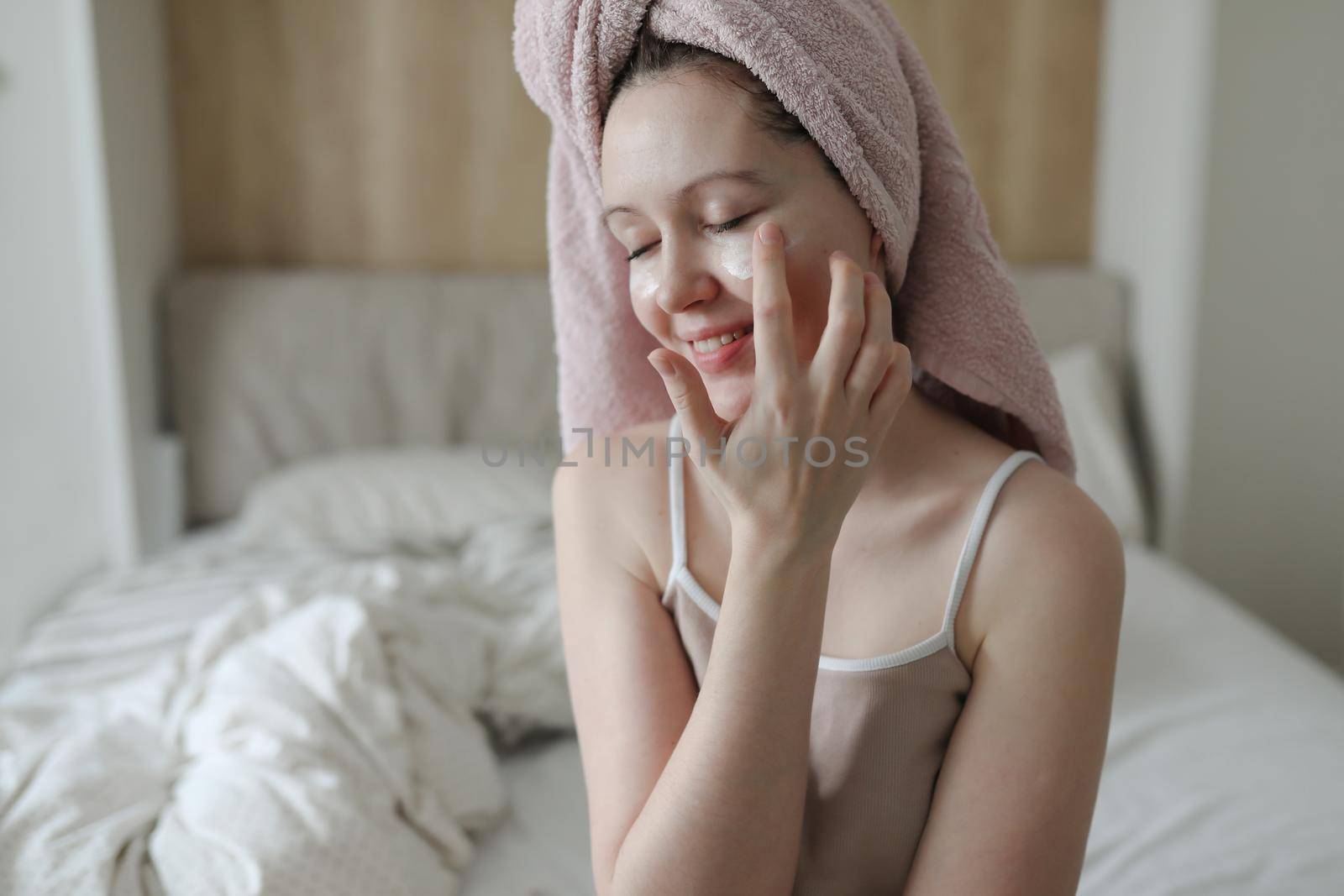 smiling young woman with a towel on head apply cream on face skin