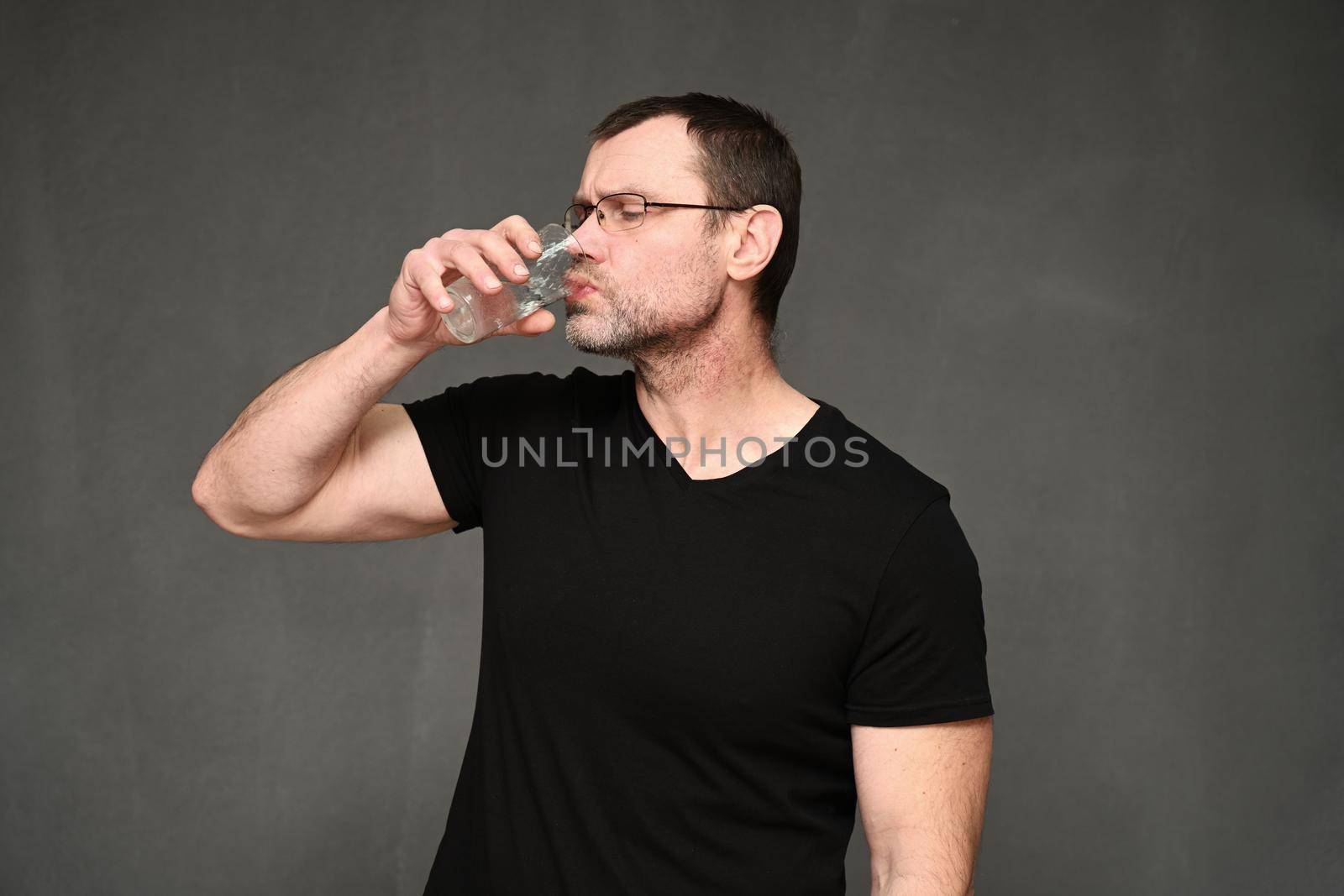 Middle-aged man in a black T-shirt and glasses drinking water from a glass by chichaevstudio
