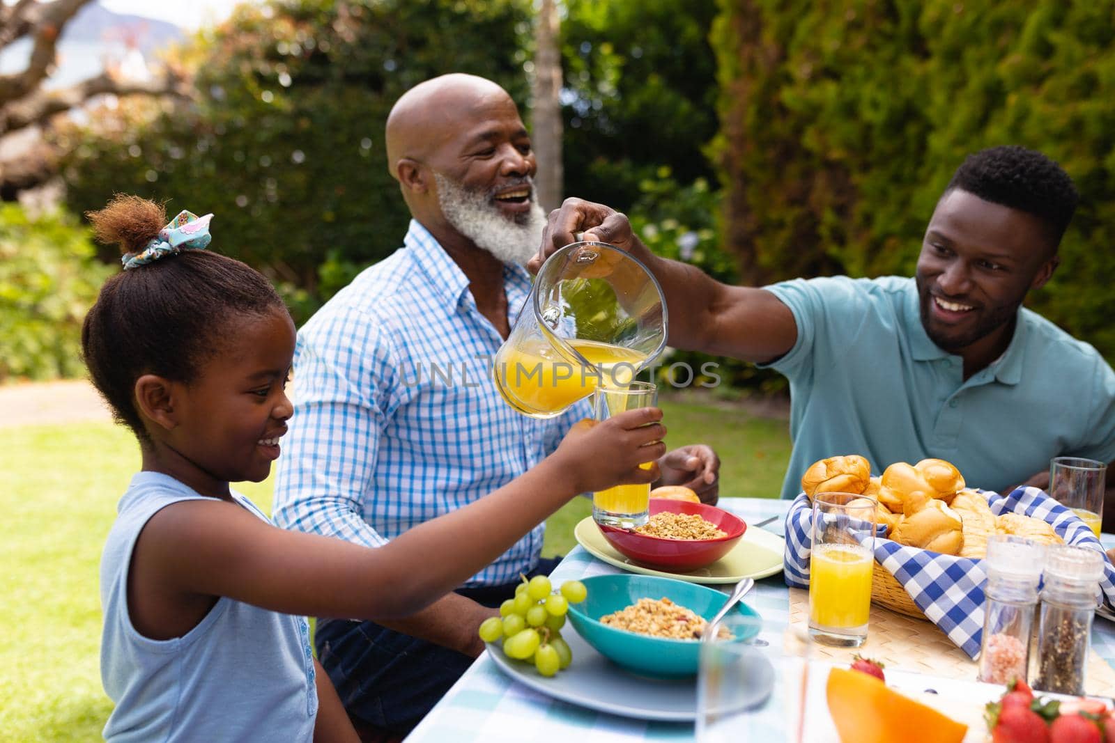 Smiling african american man serving juice for daughter by senior man at dining table in backyard. family, love and togetherness concept, unaltered.