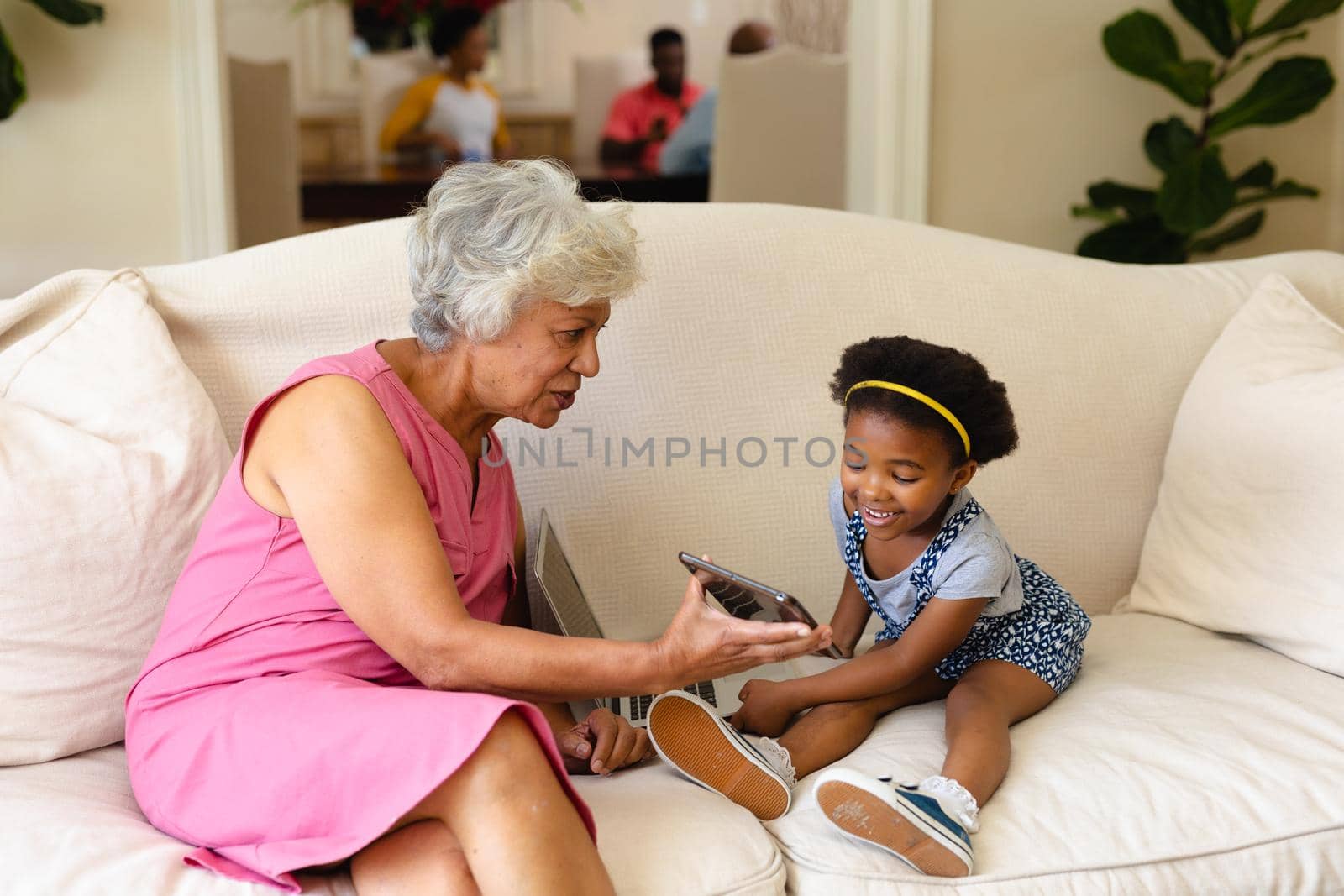 African american grandmother showing digital tablet to her granddaughter sitting on couch at home. family, love and togetherness concept, unaltered.