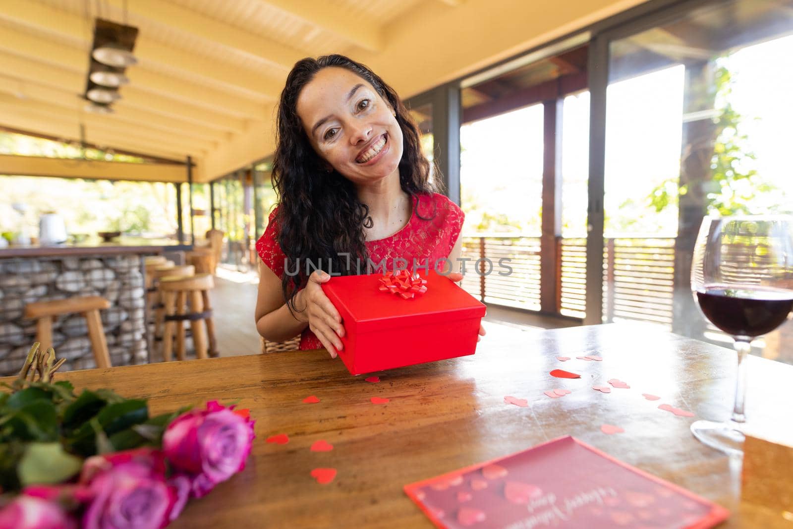 Portrait of happy biracial young woman with gift box at table in restaurant. unaltered, online dating, vitiligo and distant valentine day celebration.