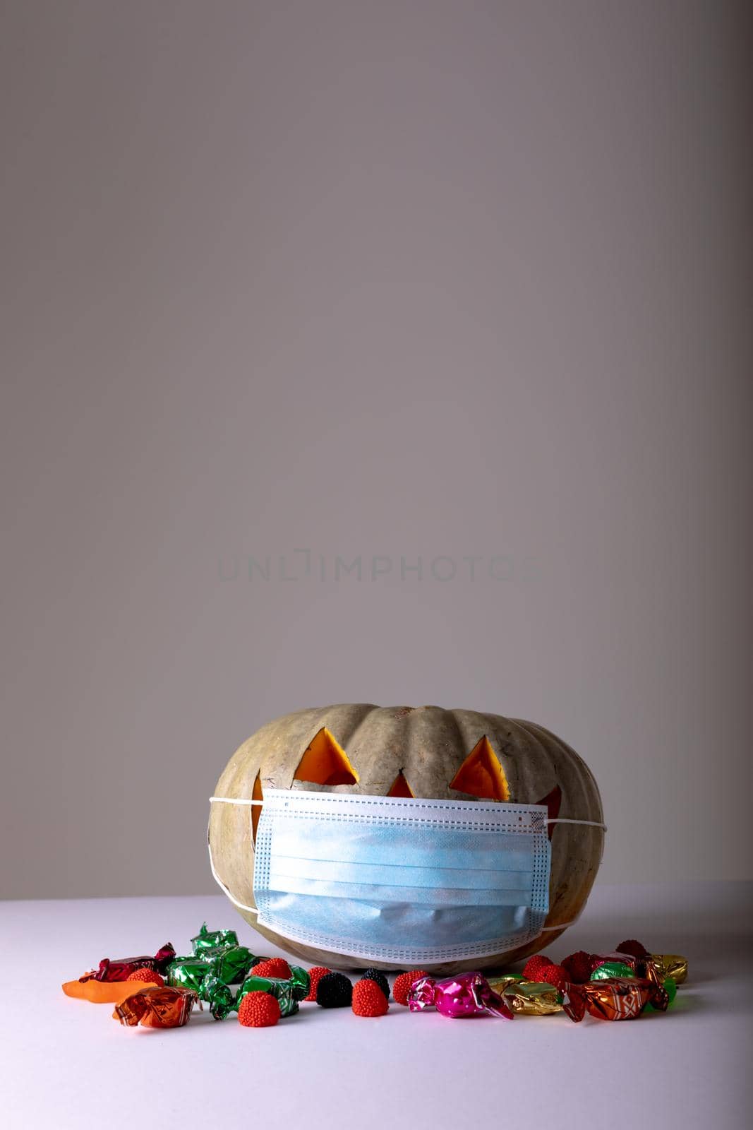 Halloween jack o lantern with face mask on grey background with sweets and copy space. halloween tradition and celebration during covid 19 pandemic concept digitally generated image.