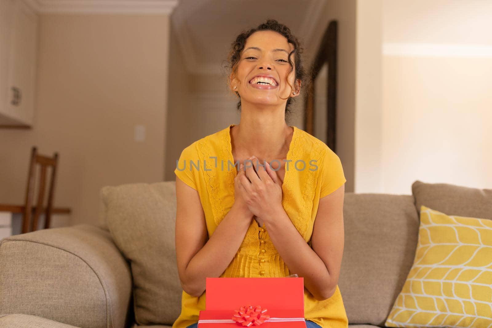 Portrait of happy young biracial woman sitting with valentine's present box on sofa at home. lifestyle and surprise, unaltered.