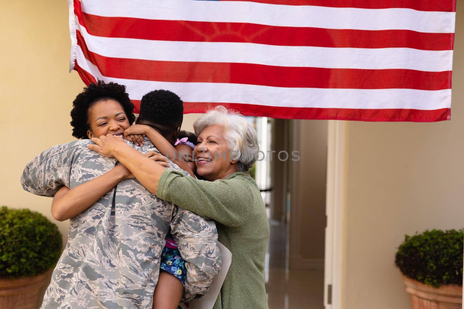 Happy african american family embracing military soldier's return at entrance of house with flag. family, bonding and patriotism, unaltered.