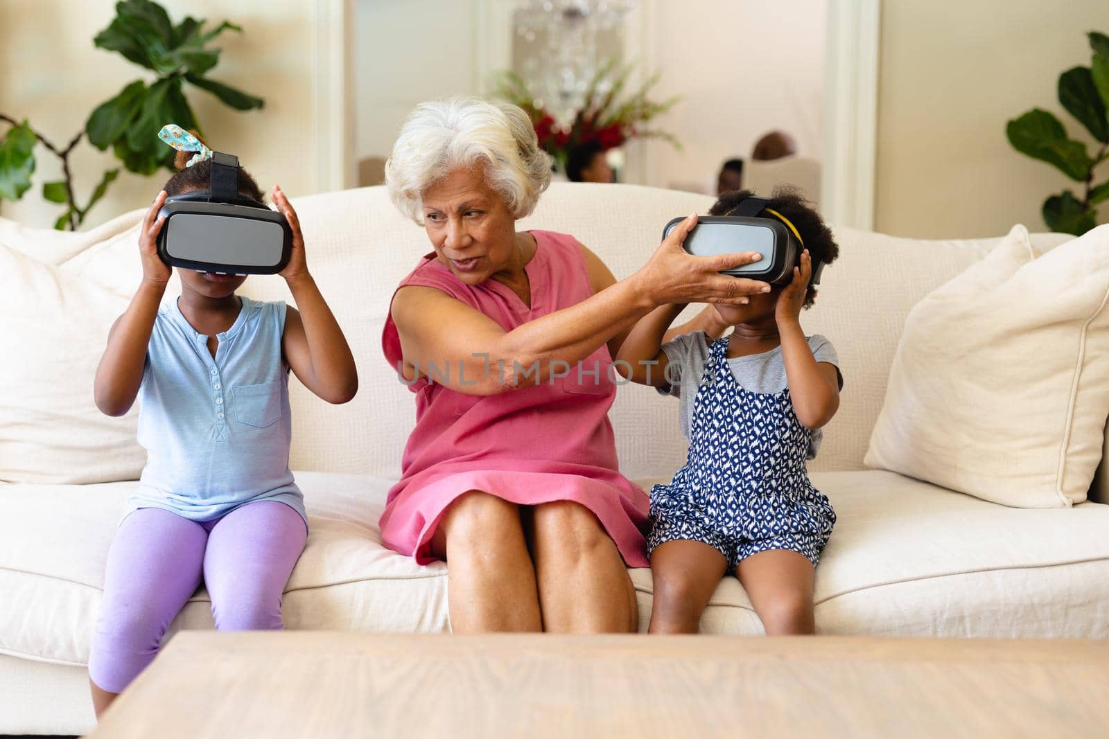 African american grandmother making her granddaughters wearing vr headsets sitting on couch at home. virtual reality and futuristic technology concept, unaltered.
