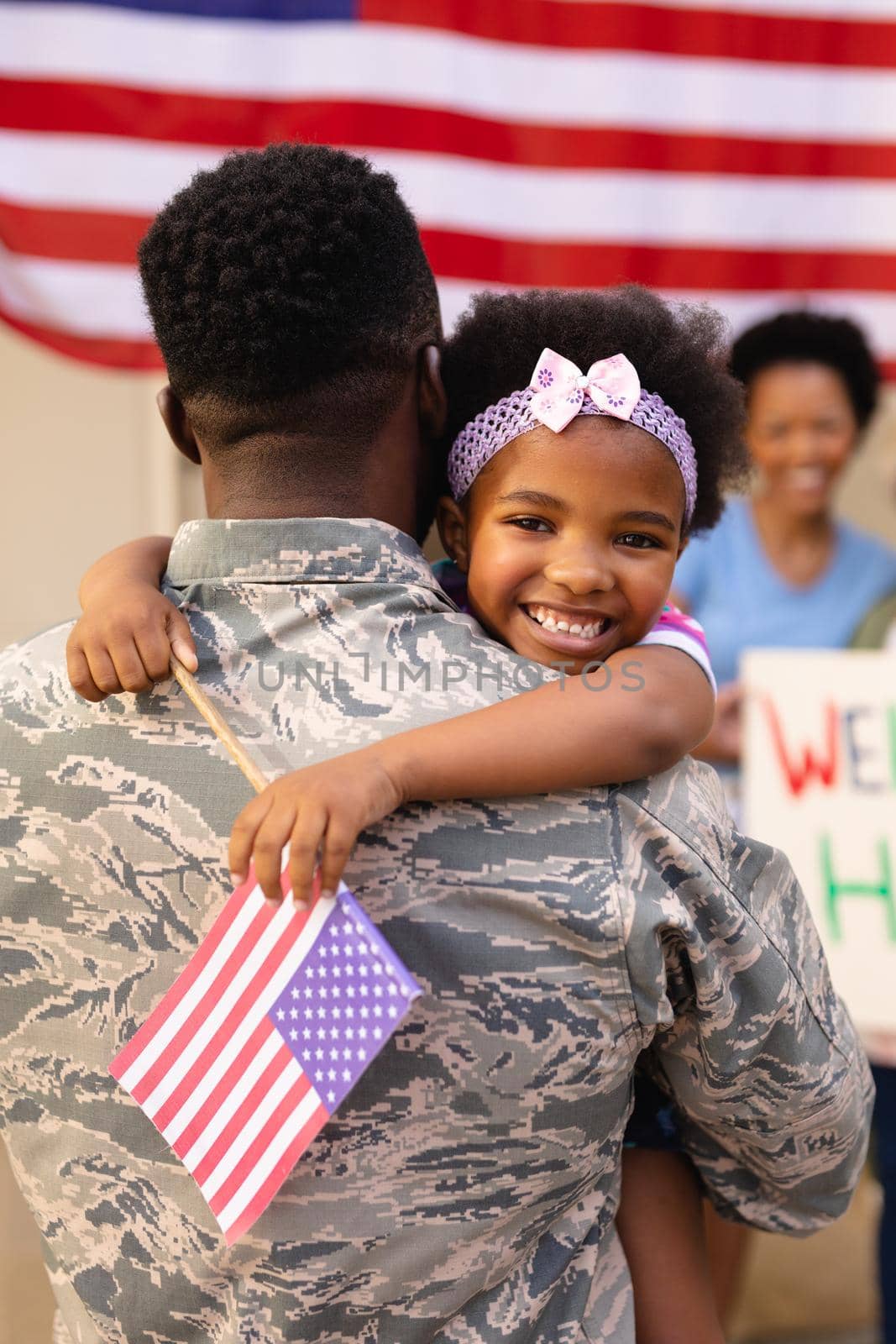 Portrait of smiling african american girl holding usa flag embracing army father on his return. family, bonding and patriotism, unaltered.